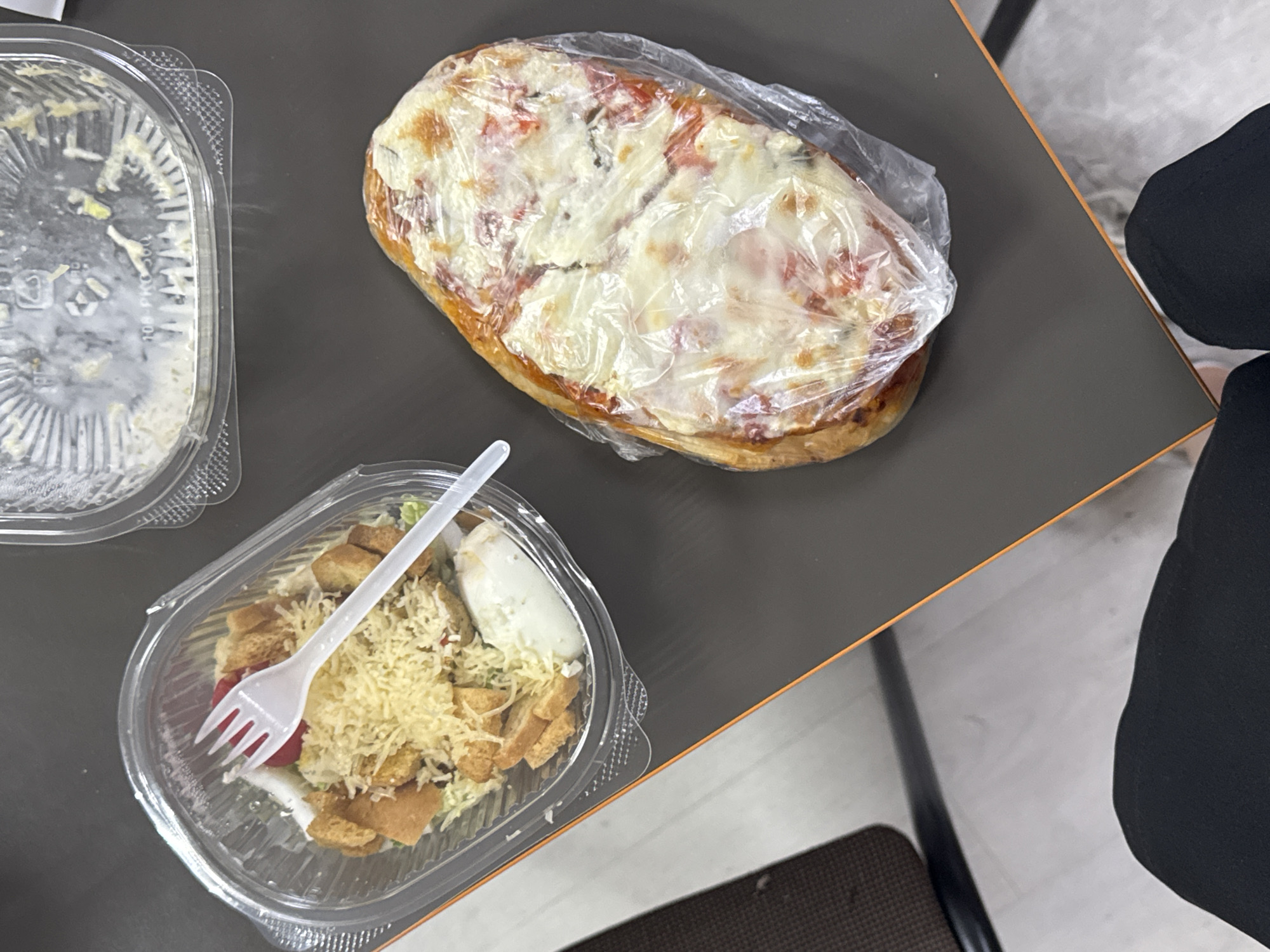 The image shows a tabletop with three plastic food containers. One container, situated in the upper-left corner, is mostly empty. Another container holds a salad with croutons, cheese, dressing, and a plastic fork. The third container holds a rectangular pizza wrapped in plastic. An individual wearing black pants sits at the table. The location is not apparent in the image.