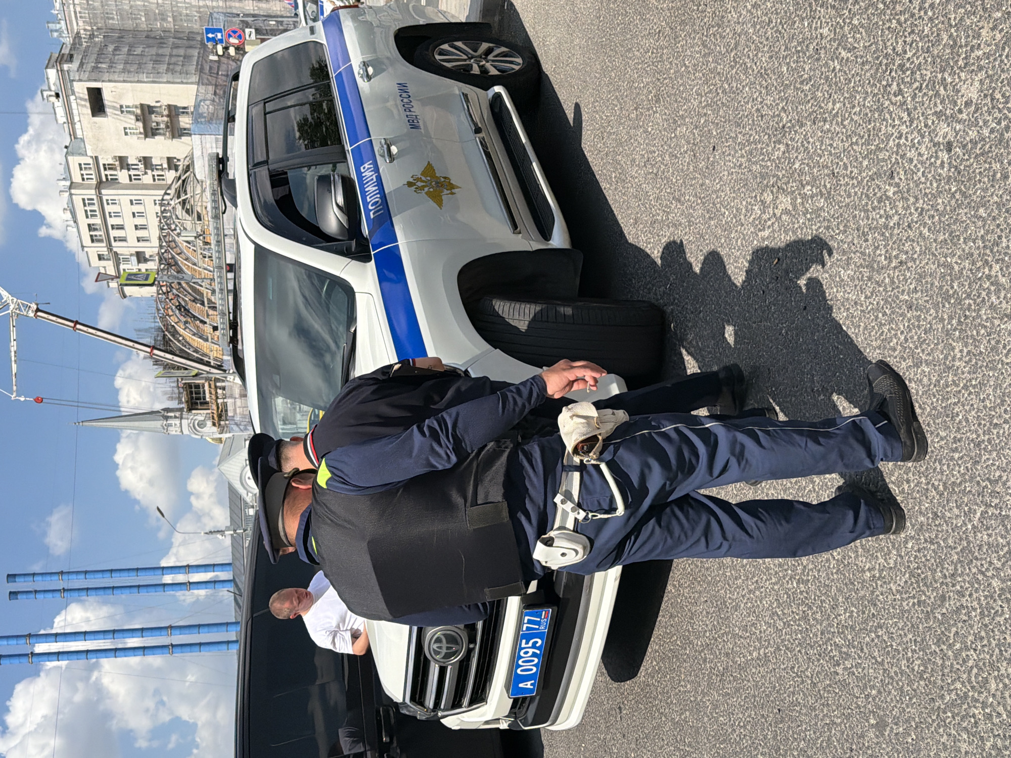In Moscow, Russia, under a clear blue sky with scattered clouds, two police officers are observed near a white police SUV. The SUV has a blue stripe along its side with "ПОЛИЦИЯ" (POLITSIYA) and "МВД РОССИИ" (MVD ROSSII) written on it, along with the emblem of the Russian Ministry of Internal Affairs. The license plate on the front of the SUV reads "А 0095 77 RUS".
One officer, facing away from the camera, wears a black tactical vest over a dark blue uniform. He has a utility belt equipped with a holster and other gear. His dark blue trousers are visible, as are his black shoes. The second officer, also in uniform, is partially visible behind the first, wearing a dark shirt and a cap.
A civilian, wearing a white t-shirt, stands near the front of the SUV, looking towards the officers. To the left, a black vehicle is partially visible. The background shows a city skyline with several tall, blue industrial smokestacks, a church spire, and modern buildings, one of which is under construction with scaffolding. A crane is also visible. The scene appears to be on a paved city street.
