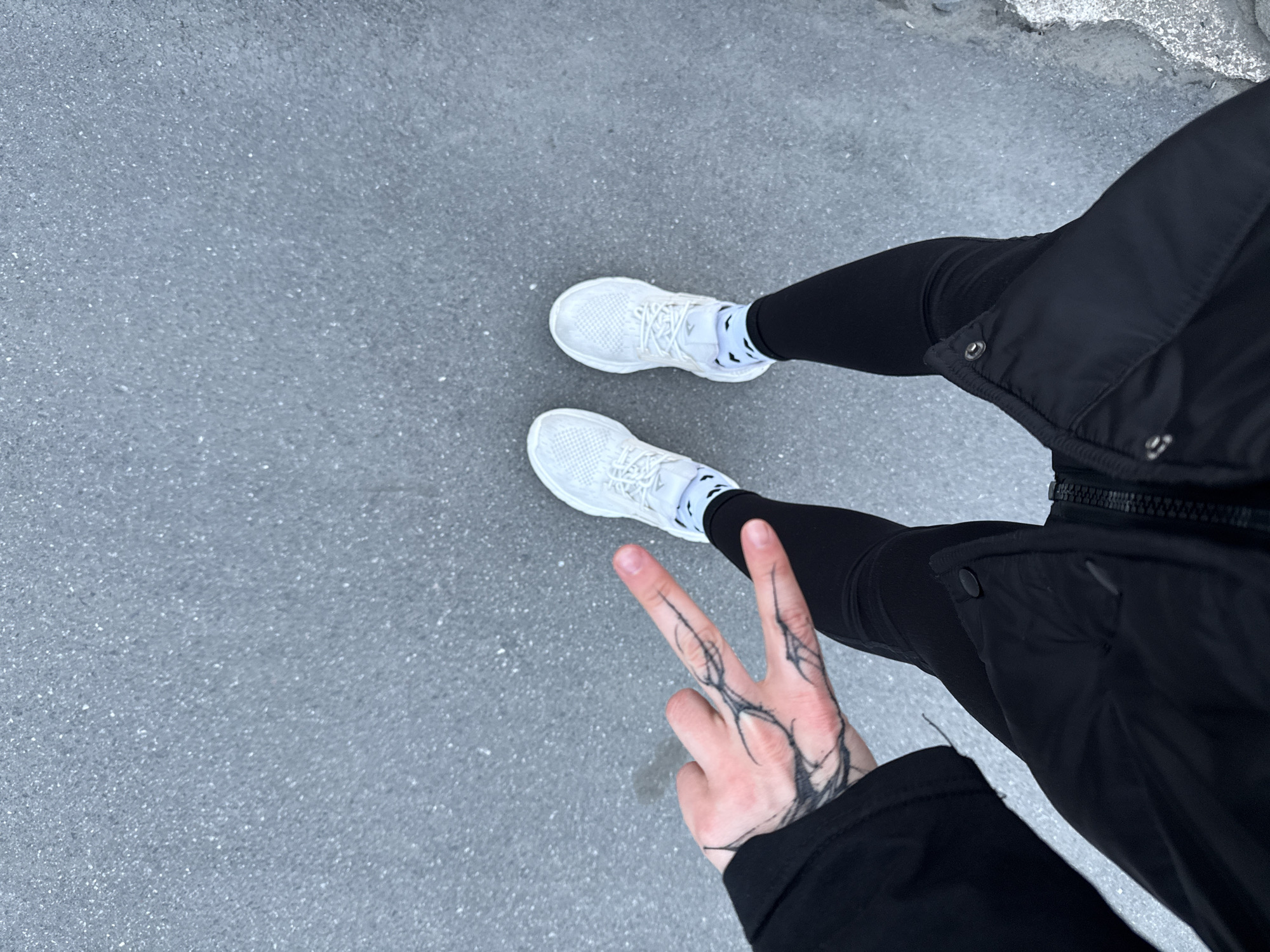 The image captures a person from a top-down perspective, focusing on their lower body and left hand. The subject is wearing black leggings, a black long-sleeved jacket, and white athletic sneakers with white laces. White ankle socks with a black pattern are visible between the sneakers and leggings.
The most notable detail is a prominent black tattoo on the back of the visible left hand, which is making a peace sign (V-sign). The tattoo features abstract, branching lines resembling veins or organic thorny structures, extending across the knuckles and fingers.
The setting appears to be outdoors on a textured, granular grey surface, likely asphalt or concrete pavement. In the upper right corner, there is a lighter, rougher patch of ground, possibly indicating ice, snow, or coarse concrete. The lighting suggests daytime. The substantial jacket and potential ground conditions are consistent with a colder climate. No discernible text is visible on any of the items.