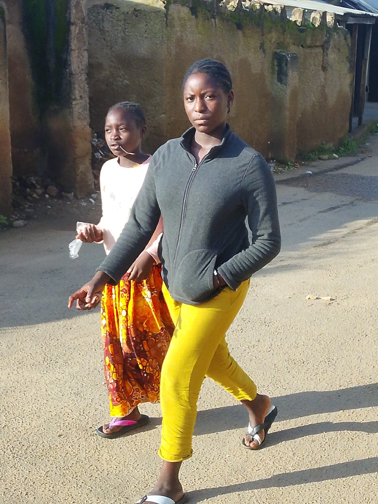 Two young females are walking on a paved surface in front of a textured, stone building. The female in the foreground, on the right, is wearing a dark grey zip-up jacket over bright yellow leggings and dark flip-flops. She is looking towards the viewer with braided hair. The female in the background, on the left, is wearing a light-colored shirt and a vibrantly patterned orange and yellow skirt with pink flip-flops. She is holding a small, transparent plastic wrapper in her right hand and looking to her left. The building behind them appears aged, with patches of moss or algae on its surface. The ground is a coarse, light-colored asphalt. The image appears to have been taken outdoors in natural daylight, casting distinct shadows on the ground. This scene is consistent with a street in the city of Jos, Nigeria.