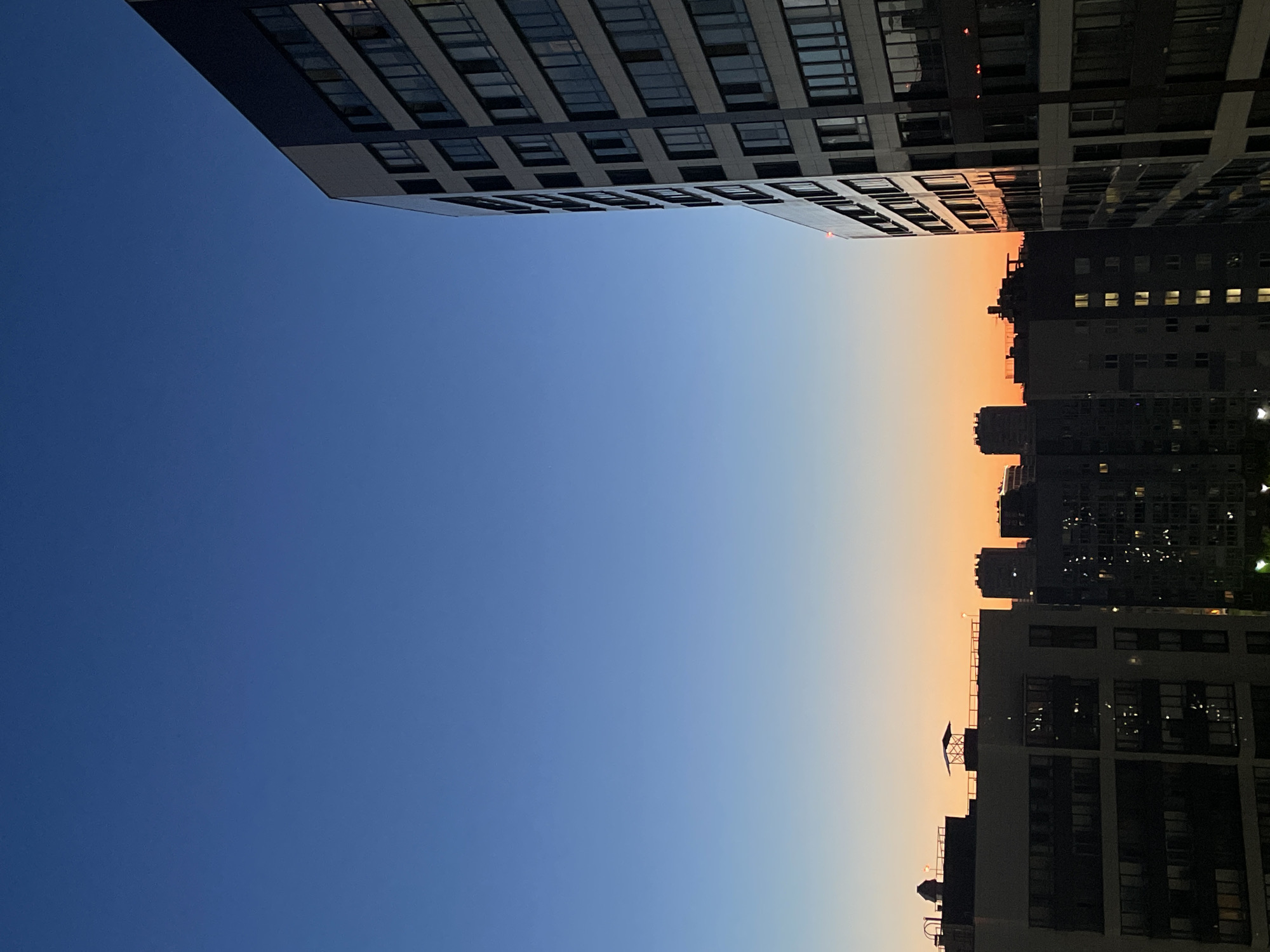 The image captures a cityscape of Sinyavino, Russia, during twilight. A tall, modern apartment building with a dark facade and numerous windows dominates the right side of the frame, extending upwards and away from the viewer. To the left and in the mid-ground, several other apartment buildings of varying heights are visible. These buildings are silhouetted against the vibrant sky, with many windows illuminated, suggesting the presence of residents. The sky transitions from a deep blue at the top to a warm orange and yellow gradient near the horizon, characteristic of a sunset or sunrise. Rooftop structures, including ventilation shafts and possibly an antenna or a small gazebo-like structure, are discernible on some of the buildings. The scene is devoid of any people or discernible activity, presenting a quiet, urban landscape bathed in the fading light.