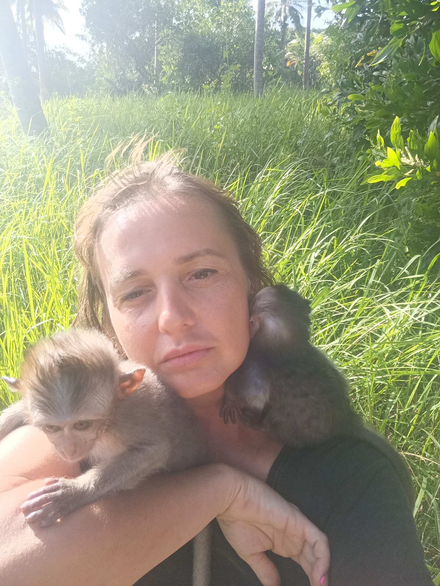 The image shows a woman in an outdoor setting, likely a park or wooded area based on the presence of tall grass, trees, and foliage in the background. The location is specified as "Unknown, Unknown." The woman is fair-skinned with light brown hair and is facing the camera. She has two monkeys clinging to her; one rests on her left arm and shoulder, while the other is situated on her right shoulder. Both monkeys are young, with one having lighter fur and the other darker. The overall lighting appears bright, possibly taken during daytime under sunlight. The woman and monkeys are the primary focus. There is no indication of urban infrastructure or identifiable landmarks to further pinpoint the location.