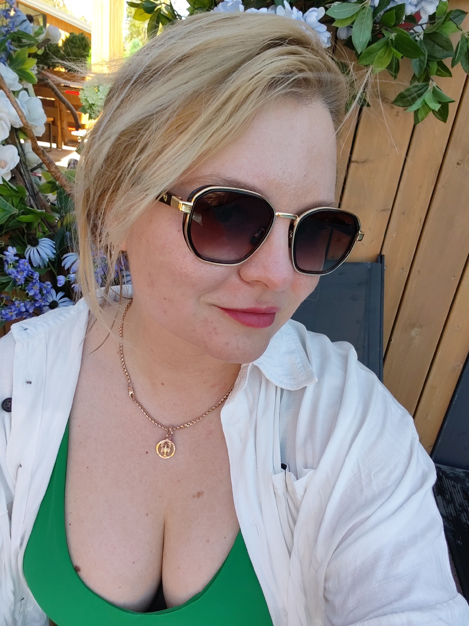 A woman with fair skin and blonde hair is posing for a picture. She is wearing sunglasses, a gold necklace, a white shirt, and a green top. The background includes blue and white flowers, greenery, and a wooden structure. The image does not contain information about the location being in the city of Izhevsk, Russia.