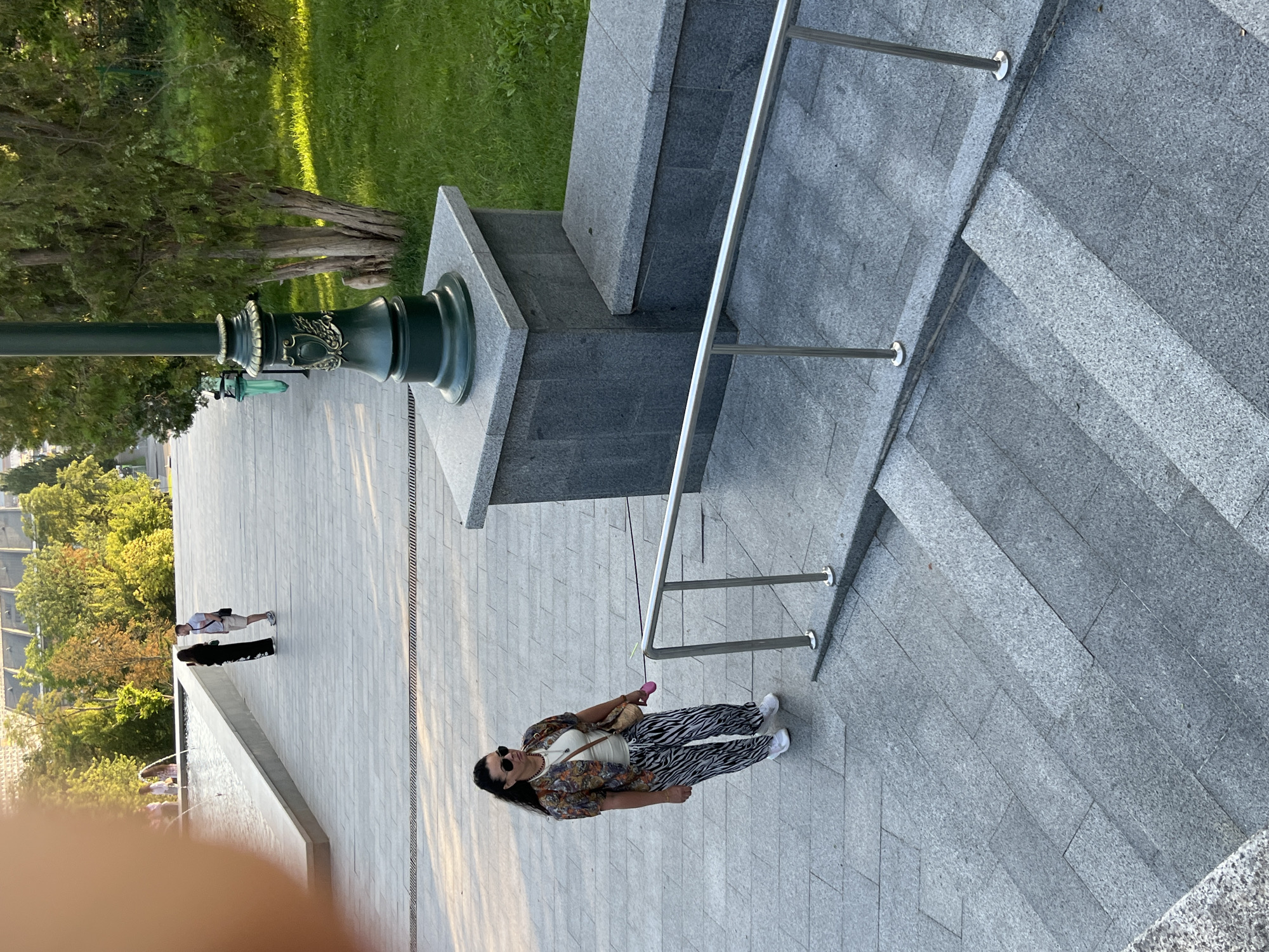 In an outdoor urban setting, a woman with dark hair and sunglasses stands on a wide, gray, paved walkway. She is wearing a patterned short-sleeved jacket over a white top and zebra-print pants, paired with white sneakers. In her right hand, she holds a pink object, possibly a toy or accessory. She is looking to her left. Behind her, a man in a white t-shirt and shorts stands by a decorative lamppost. Further in the background, two people are visible near a water feature, and a grassy area with trees borders the walkway. The scene is illuminated by sunlight, casting shadows.