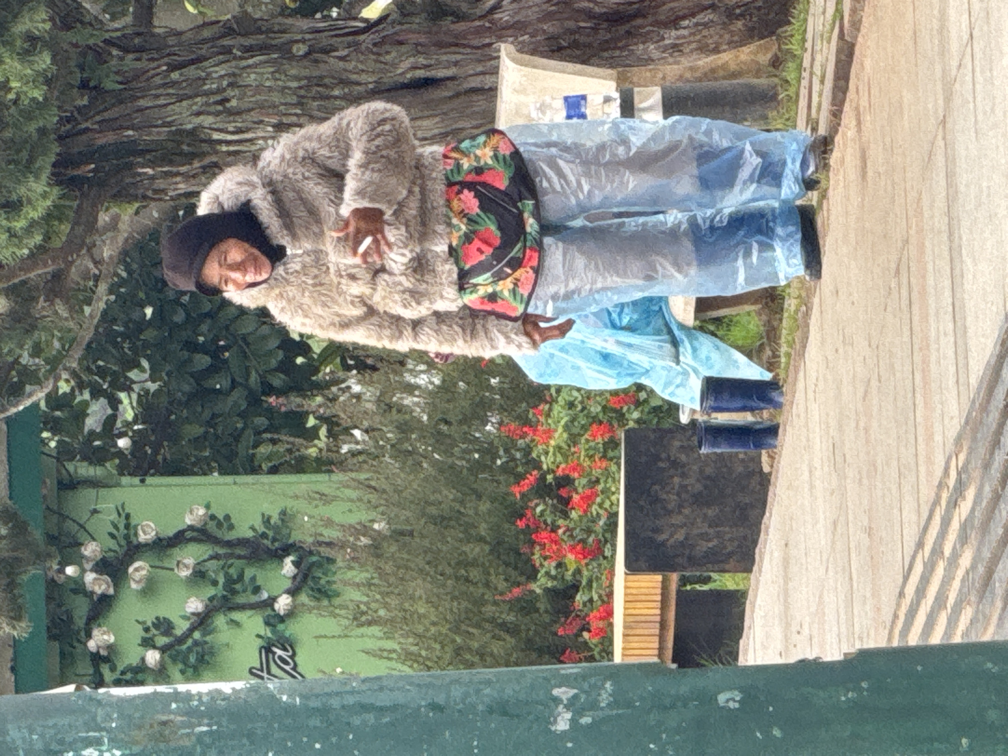 A middle-aged woman, wearing a dark headscarf, a shaggy, beige fur-lined jacket, and clear plastic pants over a floral-patterned fanny pack, stands on a paved walkway. She is holding a cigarette. Behind her, a green wall with a painted rose vine mural is visible, along with dense green foliage and red flowers. A wooden bench and dark rectangular structure are also present. In the background, a large tree with textured bark stands tall. Two dark blue cylindrical objects, possibly small trash bins or rain boots, are placed on the walkway near her. The scene appears to be outdoors, in what is identified as Da Lat, Vietnam, a city known for its cool climate and lush scenery.