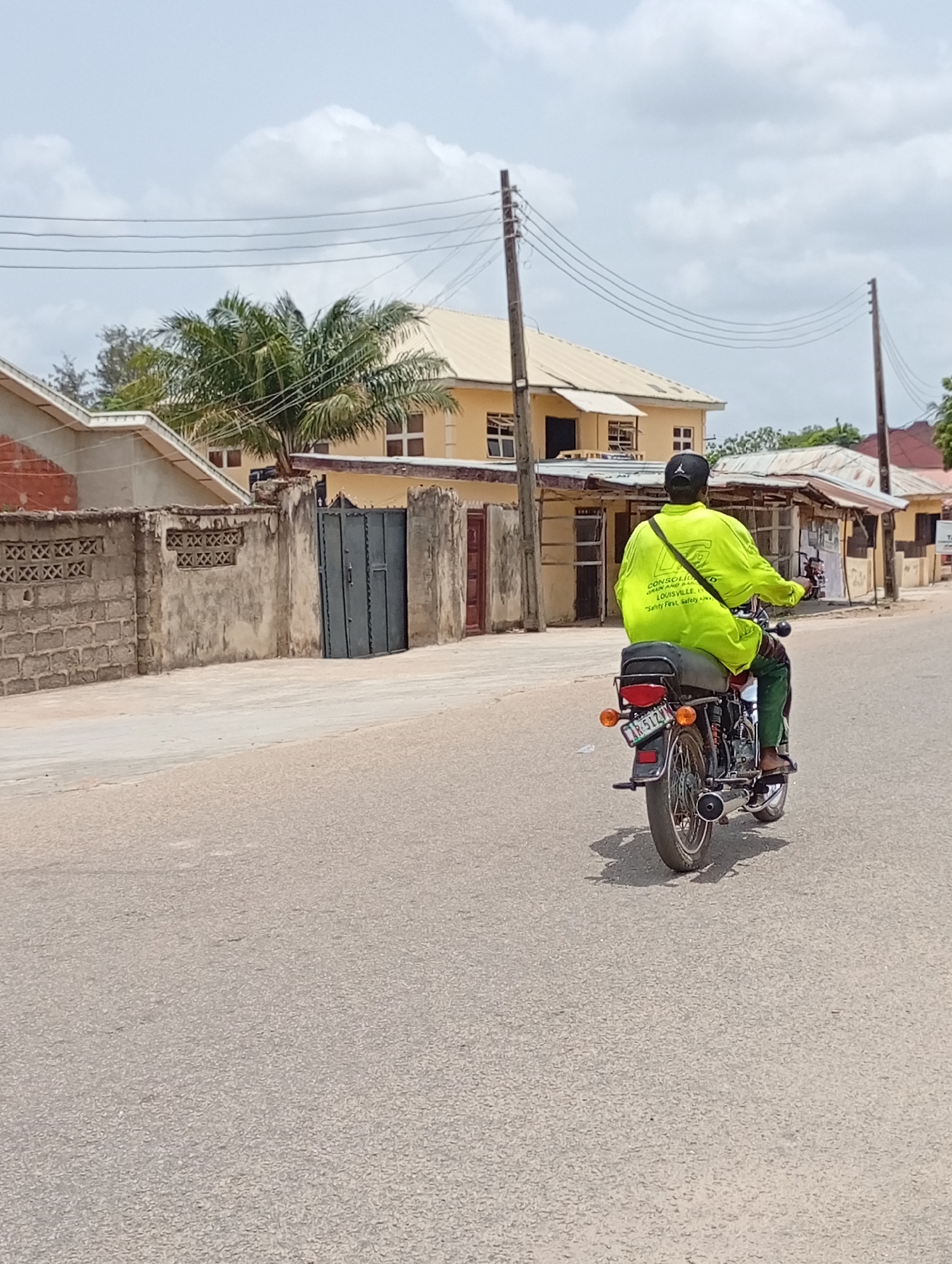 A person wearing a neon yellow long-sleeved shirt, dark pants, and a black baseball cap is riding a motorcycle on an asphalt road in Lokoja, Nigeria. The motorcycle has a license plate marked "KN-512VN" and features the text "CONSOLIDATED BRAUN AND BAR, LOUISVILLE, "Safety First, Safety Always" on the rider's back. The person is facing away from the camera. The location is a street with single-story and two-story buildings on either side, some with fences and gates. Utility poles and overhead wires are visible, and a palm tree is present in the background. The sky is overcast with some clouds.