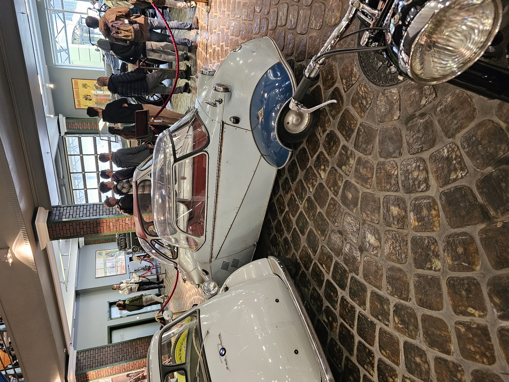 The image shows an indoor exhibit of vintage vehicles, possibly within a museum or showroom. A crowd of people is visible, observing the display. The foreground features a light-colored vehicle with a distinct design, and a motorcycle is partially visible on the right. The ground is paved with cobblestones. A second car, white in color, is partially visible in the lower center of the image. The display is secured by a red rope. A poster is on the wall in the background. Based on visual cues, the location appears to be the Архангельское, Russia area.