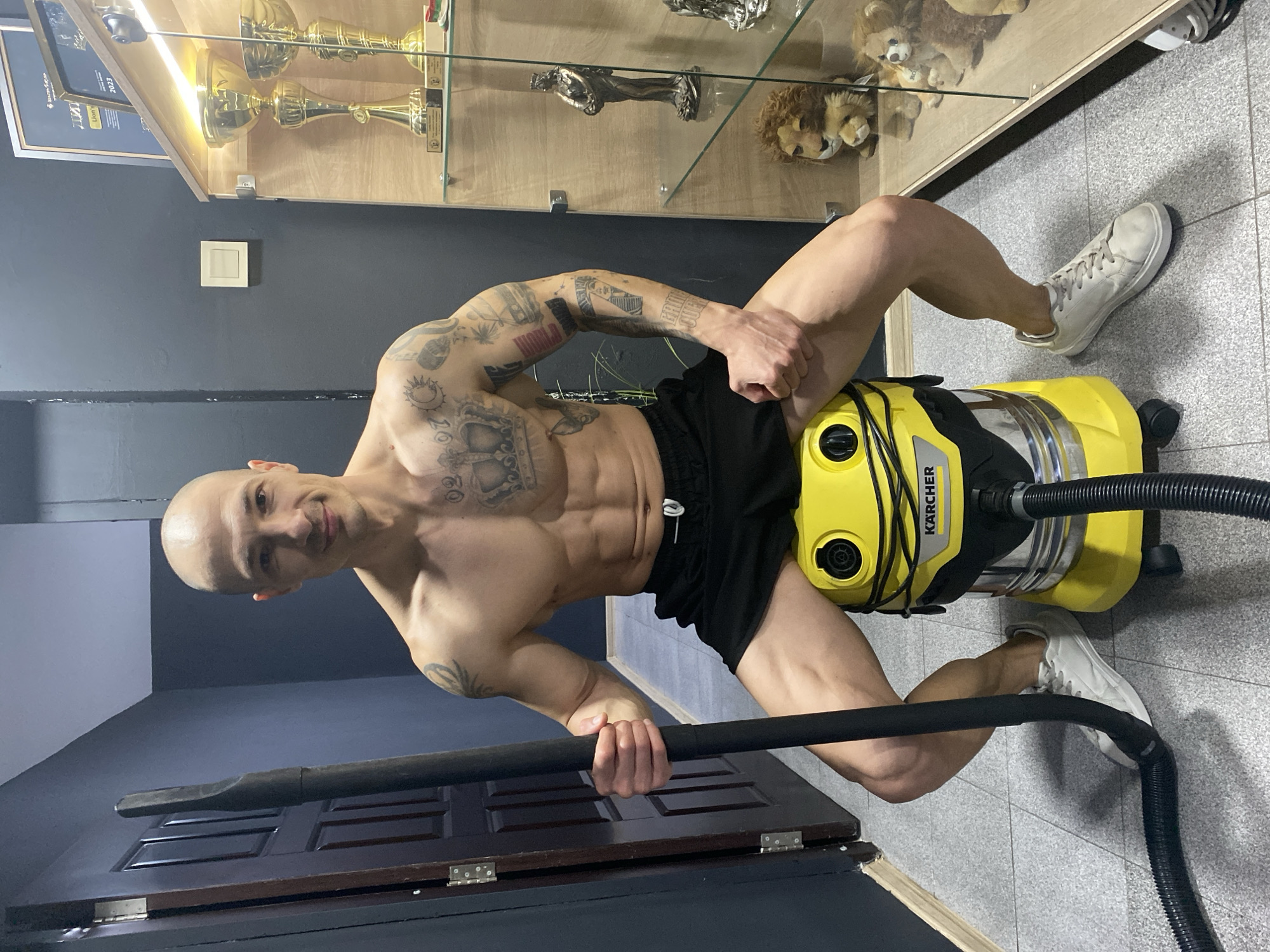 A bald, muscular male, shirtless and wearing black shorts with white sneakers, sits astride a yellow and silver Kärcher industrial wet/dry vacuum cleaner in an indoor setting in Pleven, Bulgaria. He grips a black vacuum hose and nozzle upright in his left hand. The man faces forward, looking directly at the camera with a slight smile. Numerous tattoos cover his chest and arms, including a crown design on his left pectoral and various symbols on both arms. The floor is covered with light gray tiles. To the man's left, a dark brown door with visible hinges is slightly ajar. To his right, a light-colored wooden display cabinet with glass shelves holds several gold-colored trophies, various figurines including a bronze-colored female figure and a metallic lion, and several plush toy lions. Above the display cabinet, a framed document or award, partially visible, indicates 'Lion's' and '2023'. The walls are painted a dark gray, and a light-colored rectangular light switch is mounted on the wall behind the display case.