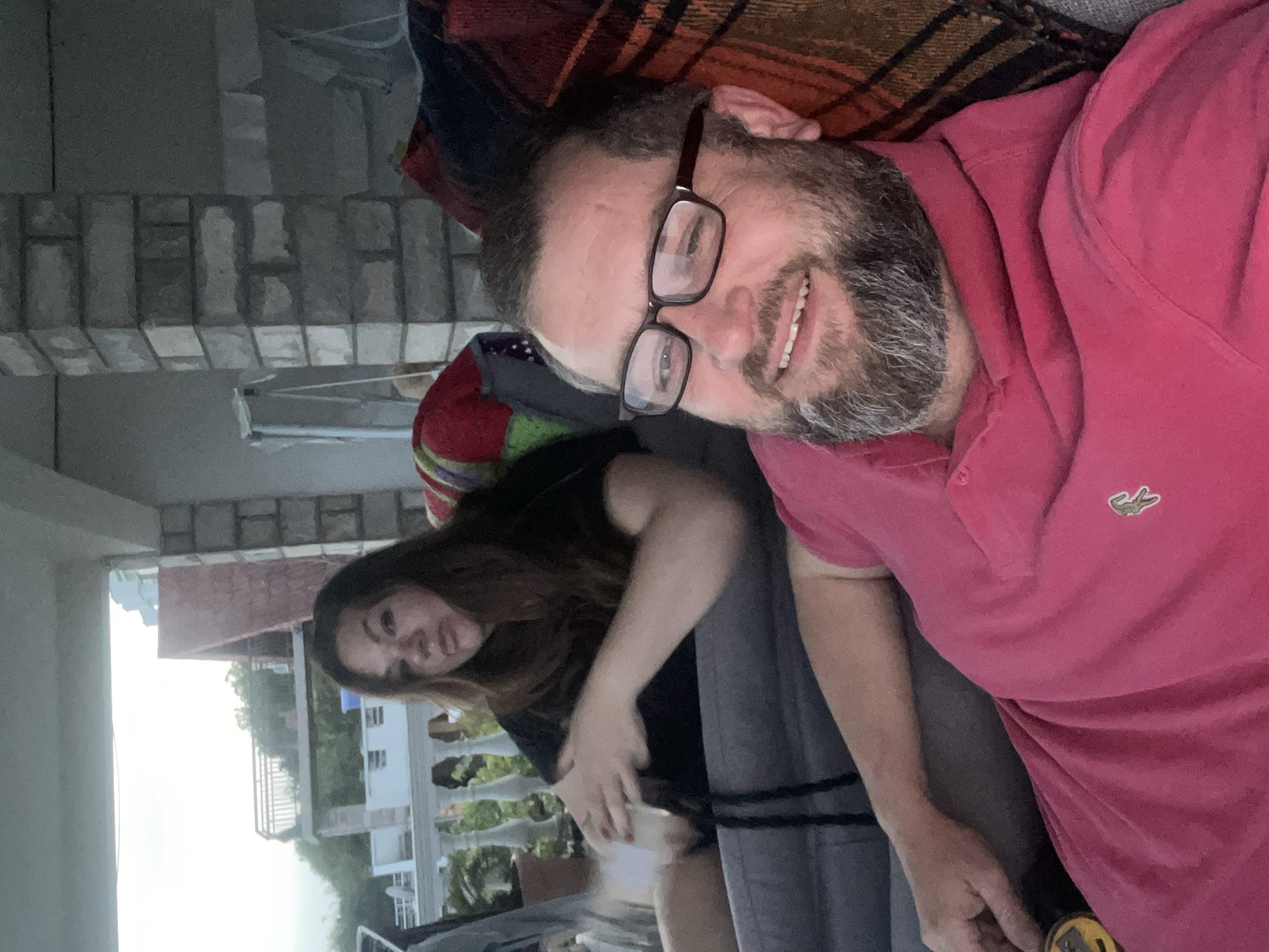 The image shows a man and woman sitting together. The man is wearing glasses and a pink Lacoste polo shirt. He has a beard and is smiling. The woman is wearing a black shirt. They are sitting on a gray couch or chair, with a patterned blanket behind the man. In the background, there is a view of a building with white balconies and possibly a red roof, along with trees and a hint of sky. The location is likely Bijela, Montenegro, based on the cityscape visible in the background.