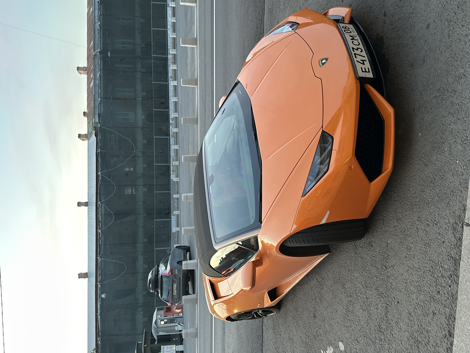 An orange Lamborghini coupe, bearing a Russian Federation license plate with registration number “E473CM 08”, is parked on an asphalt surface. In the background, a dark-colored SUV is parked near a gray building facade and a series of white barriers. The setting appears to be Saint Petersburg, Russia, based on the vehicle's license plate.