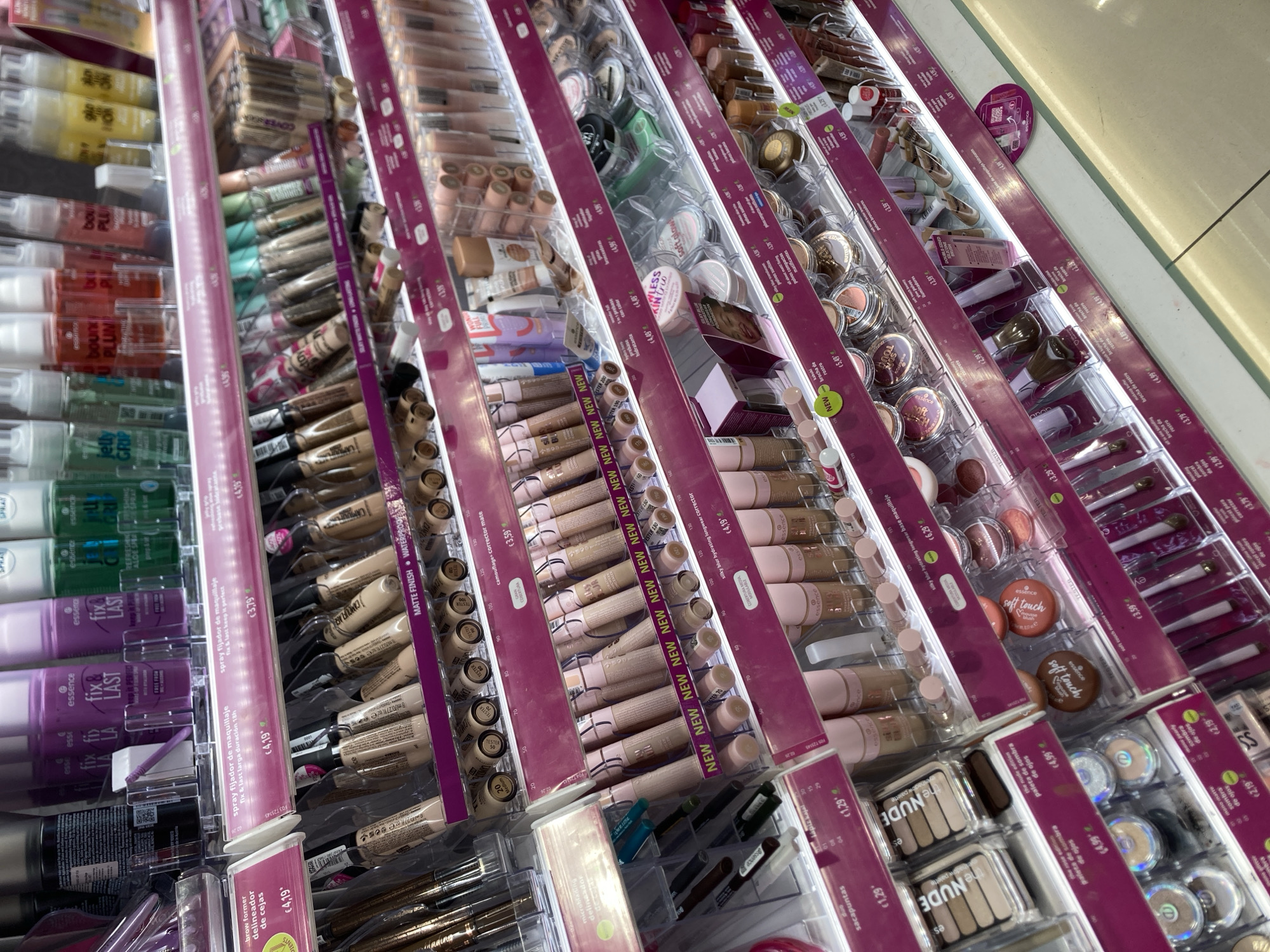 The image shows a retail display of makeup products in a store, likely a cosmetics retailer or a drugstore, given the brands and product types. The products are arranged on several shelves, with various items such as concealers, foundations, eyeshadow palettes, blushes, and makeup brushes visible. Many of the products have pink and purple branding, with the brand "essence" being prominent. Prices are displayed on small white tags in Euros, such as "€4.19" and "€3.79". Some products are labeled as "NEW". The overall lighting is bright, illuminating the colorful packaging of the makeup. The arrangement suggests a well-organized retail environment. The specific location of Barcelona, Spain is not visually evident in the image, but the presence of Euro pricing suggests a European country.