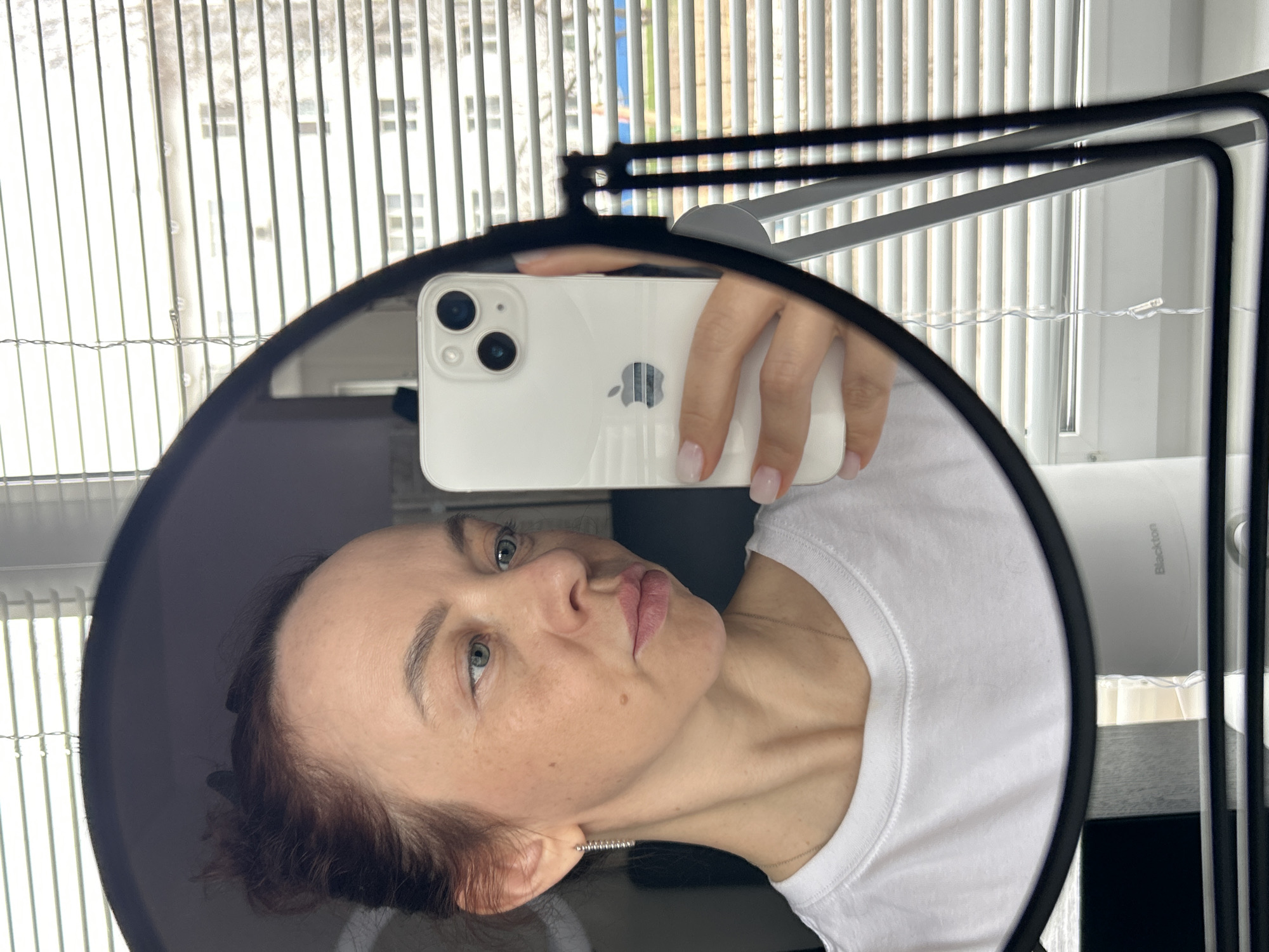 A woman is taking a selfie with a white iPhone inside a ring light. She is wearing a white t-shirt. The background shows a window with blinds and a blurred cityscape. The picture was taken indoors, likely in a residential area. It's impossible to verify the location in Chaikovsky, Russia, from this image alone.
