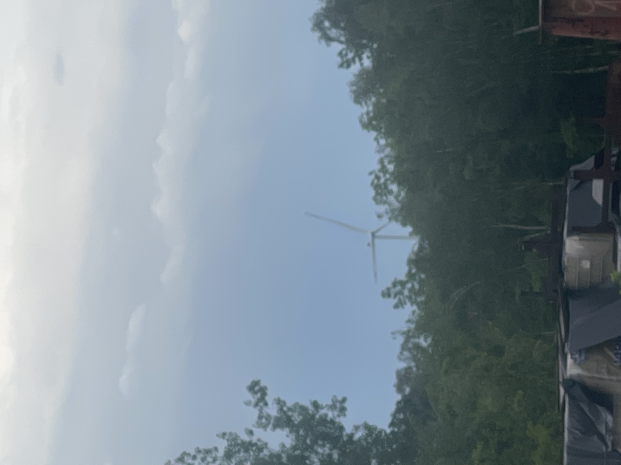 The image captures a static outdoor scene dominated by a large wind turbine visible in the distance against a cloudy, light blue and white sky. The turbine is positioned towards the center-right of the frame, with its tower and some blades distinctly silhouetted against the sky.
The right side of the image and extending towards the bottom is filled with dense green foliage, likely trees, suggesting a natural or vegetated area. On the far right edge of the frame, obscured by the trees and cut off, are parts of what appear to be structures or buildings with dark roofs and some lighter-colored elements.
The sky occupies a significant portion of the left and upper parts of the image, showing an overcast or partly cloudy condition with soft light, indicating daytime. No human figures or discernible activities are present. The overall impression is one of a serene, possibly rural or industrial-edge landscape. There is no clearly visible or decipherable text in the image.