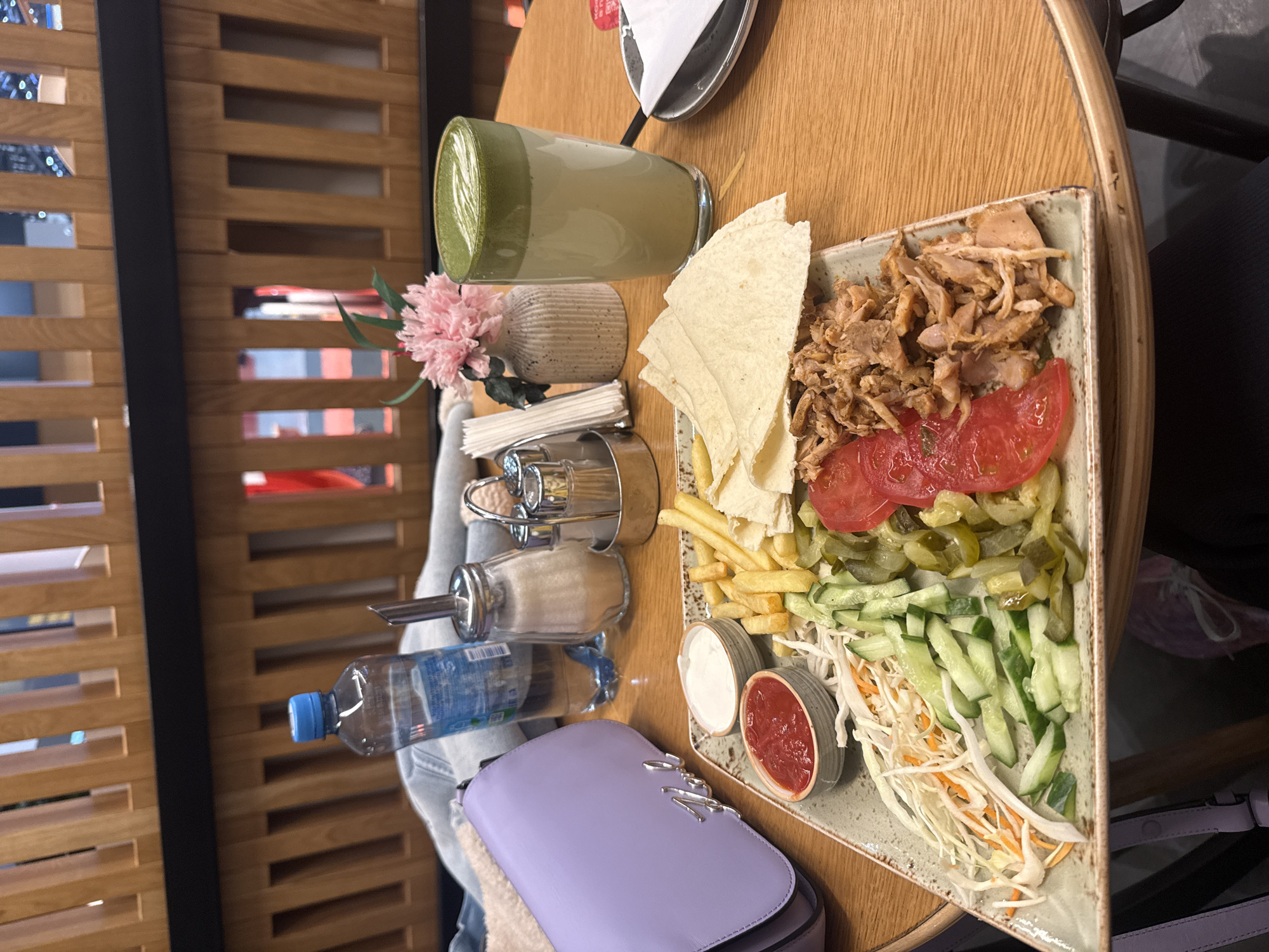 The scene depicts a meal set on a light-brown wooden table inside a cafe or restaurant in Khimki, Russia. A large rectangular plate in the foreground holds a serving of shredded cooked meat, sliced red tomatoes, sliced pickled cucumbers, sliced fresh cucumbers, shredded white cabbage and carrots, french fries, and several pieces of flatbread. Two small, ridged ceramic bowls on this plate contain red ketchup and a white sauce. To the left of the plate, a lavender-colored handbag with silver "KARL" lettering is visible, alongside a clear plastic bottle of Aqua Minerale water with a blue cap. Behind the plate, a glass sugar dispenser with a metal spout and a metal caddy containing salt and pepper shakers and white paper napkins are present. A small, ribbed vase holding a pink artificial flower sits next to a tall glass containing a green-layered beverage with foam art, consistent with a matcha latte. A small dark plate with a folded white napkin rests to the right of the latte glass. The background features a slatted wooden wall with horizontal and vertical elements and a dark horizontal beam. A leg, clad in dark fabric, is partially visible under the table on the lower-left.