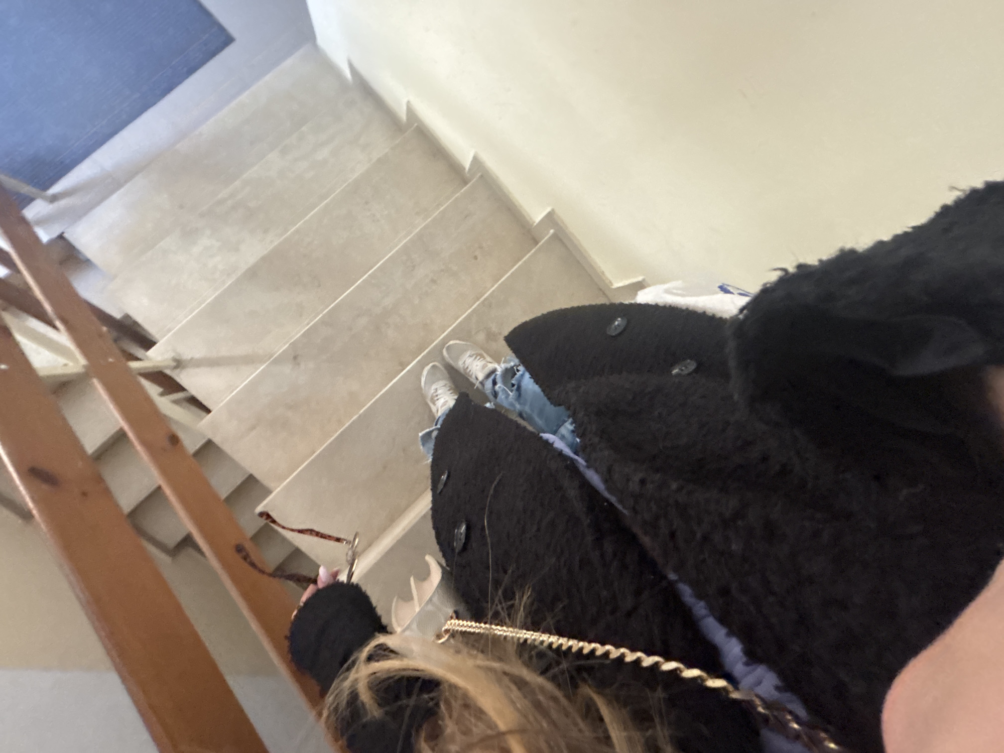 A low-angle, point-of-view shot captures a person standing on a light-colored staircase, looking down. The person's light-colored sneakers and ripped blue jeans are visible on one of the steps. They are wearing a black, fuzzy or teddy-style coat. Part of the person's blonde hair is seen in the foreground, along with a golden chain, possibly from a bag strap, and a hand holding a small item with a ring or clasp. The staircase consists of light-colored steps, possibly marble or concrete, and features a wooden banister or railing structure on the left side. The walls alongside the stairs are a plain, light beige or cream color. At the top of the stairs, a blue mat or carpet is visible. The scene depicts an indoor staircase, possibly within a residential or public building in Veliko Tarnovo, Bulgaria. There is no visible text in the image.