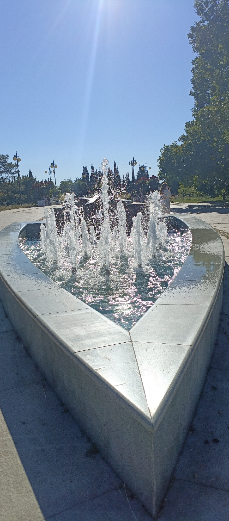 A heart-shaped fountain with multiple jets of water is centrally located in the foreground. The fountain basin is made of grey stone and is filled with water that reflects the sunlight. In the background, there is a clear blue sky with the sun shining brightly, creating lens flare. Beyond the fountain, a park-like setting is visible with trees, streetlights, and a stone monument. A woman is standing in the background, facing away from the camera, and appears to be looking at the monument or the trees. The ground surrounding the fountain is paved with grey stones.