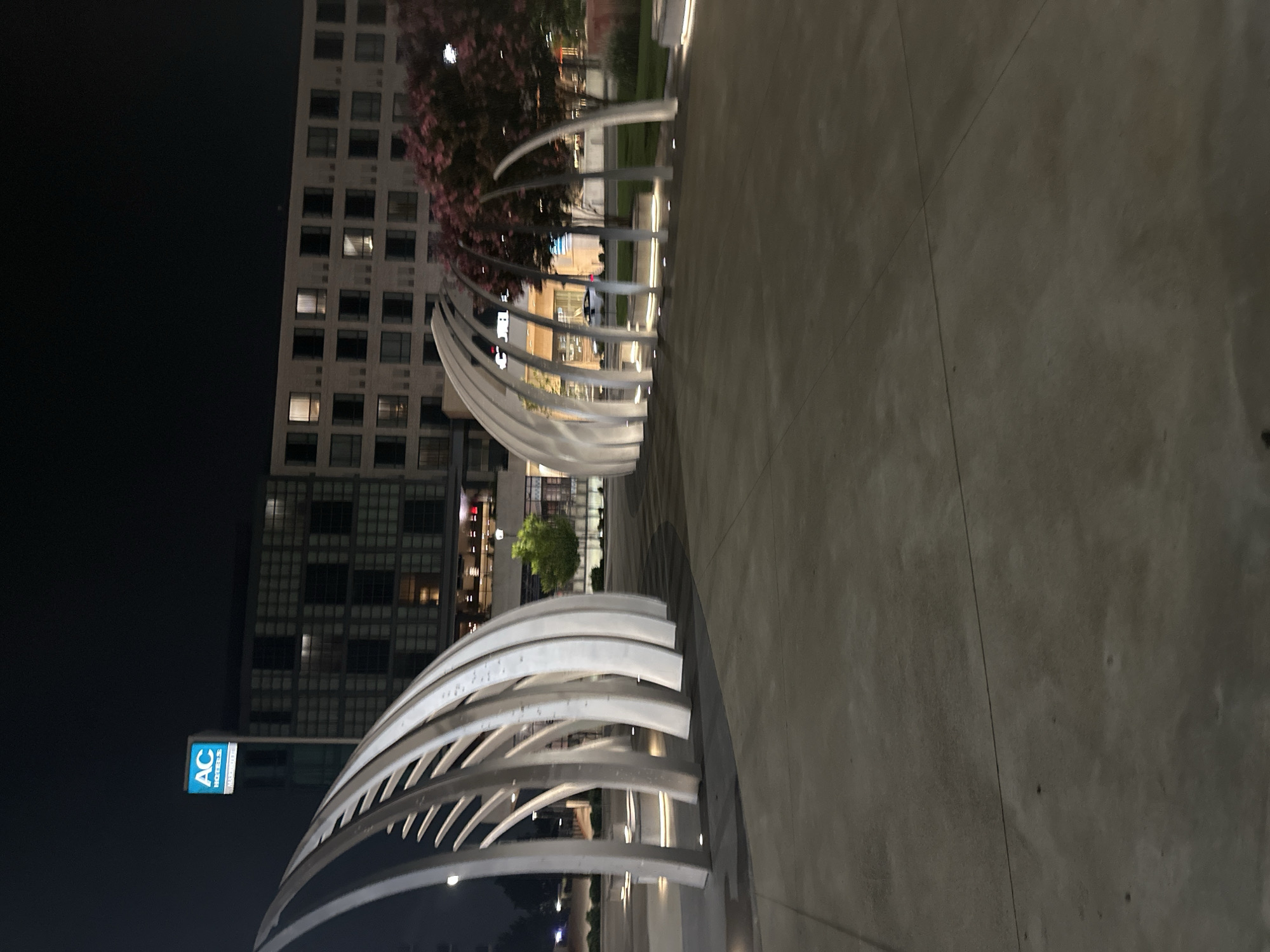 The image shows a nighttime view of an outdoor public space featuring two curved, modern art installations made of white metal. These installations are reminiscent of rib cages or arched structures and are illuminated from below by soft white lights. In the background, a multi-story building with a modern facade is visible, with many windows, some of which are lit from within. A blue and white sign for "AC HOTELS" is mounted on the building. Several trees with pink blossoms are also present in the background, along with streetlights and the general urban landscape at night. The ground in the foreground is a smooth, dark concrete surface, indicating a plaza or walkway. The location is identified as Huntsville, United States of America, suggesting this is an urban development or public area in that city. There are no people or animals visible in the image.