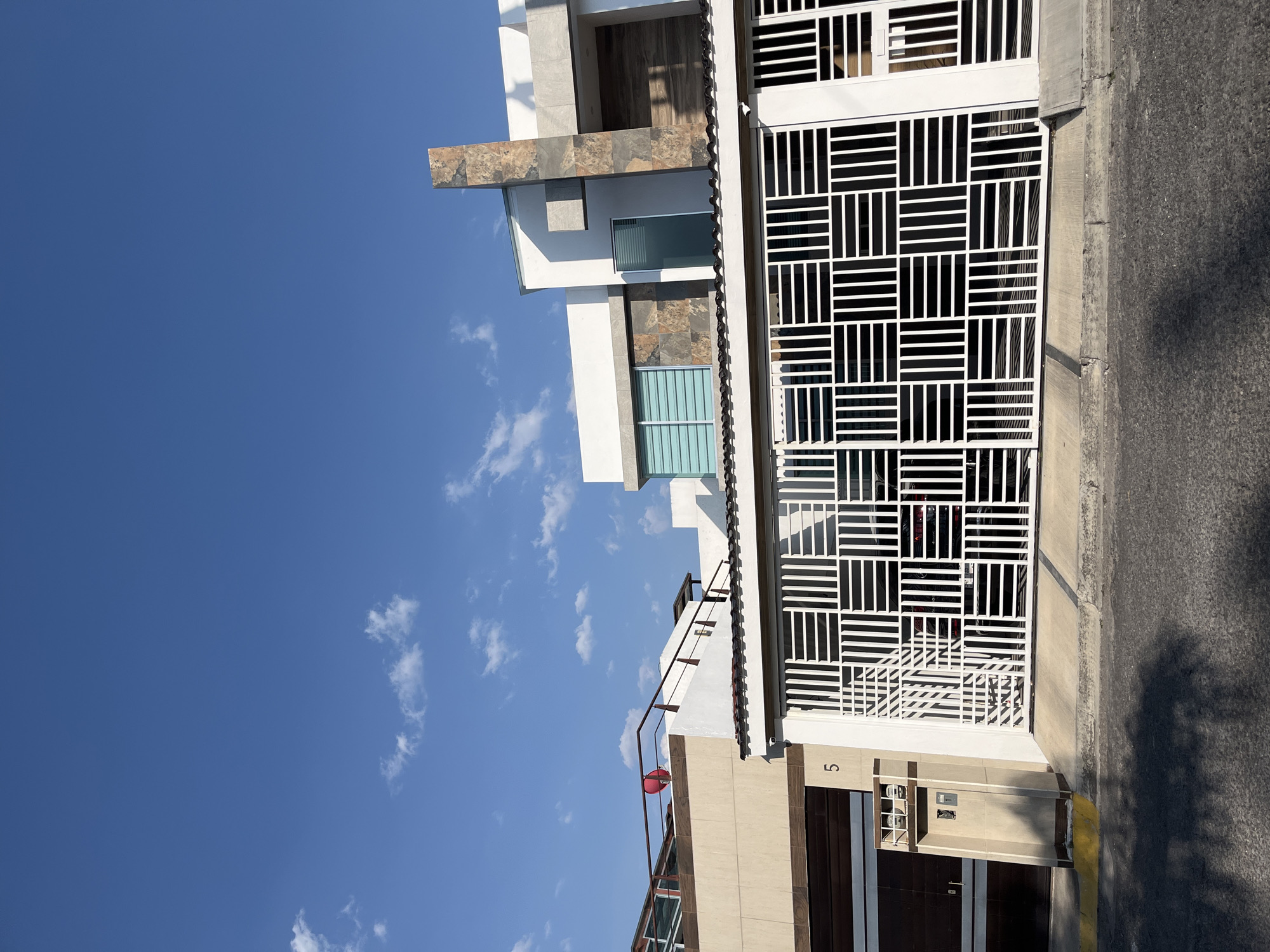 Exterior view of a modern building in San Bernardino Tlaxcalancingo, Mexico. The building features white walls, stone accents, blue-tinted windows, and a white metal gate. The gate has a grid pattern and is positioned next to a sidewalk. The number "5" is visible on the building's exterior. Above the building is a clear blue sky with scattered clouds.