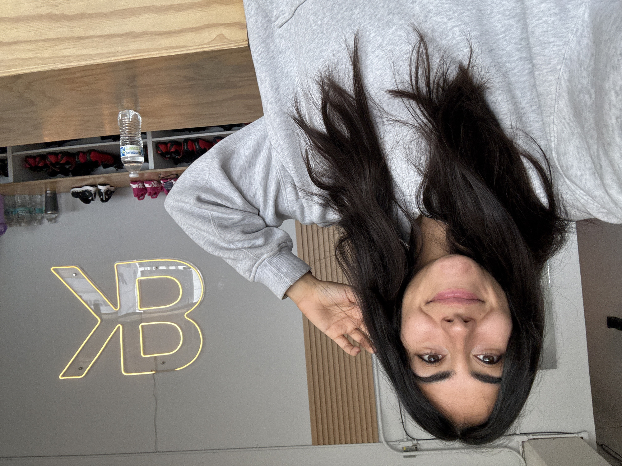 A medium shot shows a woman with long, dark hair and fair skin, wearing a gray sweatshirt. She is oriented upside down in the frame, smiling at the camera. Behind her is a light gray wall with a large neon sign in yellow, shaped like the letters "KB" intertwined. Above and behind the woman is a wooden shelf. A water bottle is visible on the shelf, alongside what appears to be an assortment of shoes. Given the architecture and interior design, the image might be from a fitness studio or gym in Puebla, Mexico. This cannot be proven forensically. The absence of precise forensic information about the Puebla location limits the analysis.