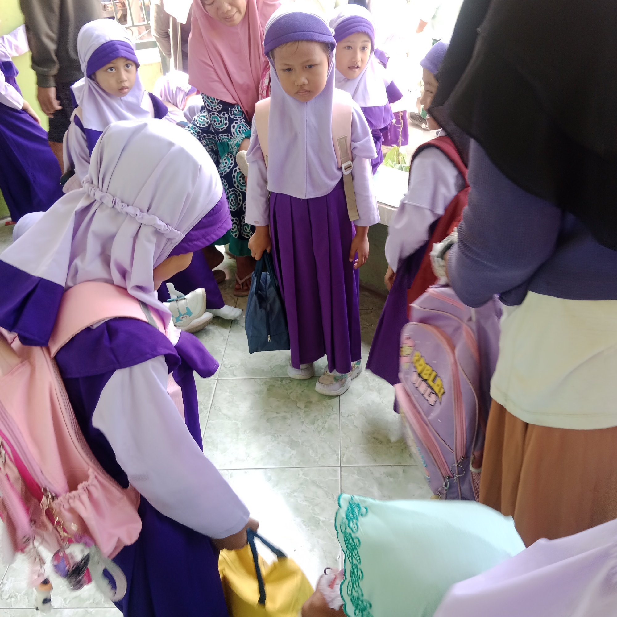 In Tegowanu Kulon, Indonesia, a group of young girls wearing purple and white hijabs and uniforms are gathered, some carrying backpacks. The floor is tiled in a light green and white checkered pattern. In the foreground, a girl in a purple hijab and uniform is being filmed, with her pink backpack visible and adorned with charms. Another girl, also in a purple hijab and uniform, stands to her right, holding a blue bag. Behind them, other girls are visible, as well as adults in the background, one wearing a pink hijab and the other a black hijab. The overall scene appears to be outside a building, possibly a school, given the context of the children's attire. The lighting suggests it is daytime.