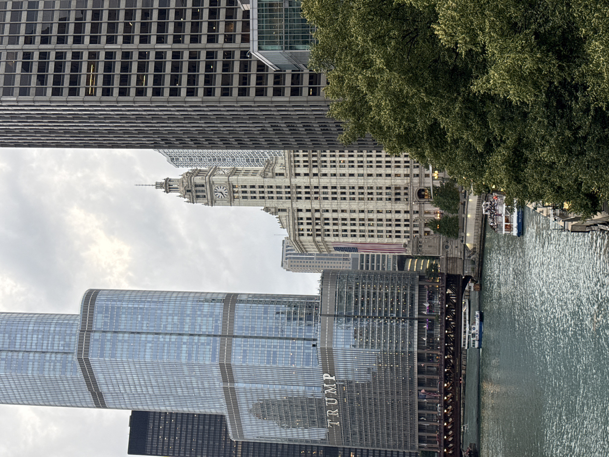 This is a medium-shot, eye-level, vertical photograph taken on an overcast day in Chicago, Illinois, United States of America. The scene captures a portion of the Chicago River with several boats on it, bordered by impressive skyscrapers and lush green trees.
Dominating the left side of the frame is the Trump International Hotel & Tower, a contemporary skyscraper with a glass facade that reflects the cloudy sky. The word "TRUMP" is prominently displayed in large, silver lettering on the lower section of the building. Adjacent to it, to the left, is a dark-colored skyscraper.
To the right of the Trump Tower, the historic Wrigley Building stands out with its ornate architecture and a prominent clock tower featuring a large clock face. An American flag is visible on the side of the Wrigley Building. Further to the right, a darker, more modern skyscraper with a grid-like pattern of windows occupies a significant portion of the frame.
In the foreground, the Chicago River flows, appearing somewhat choppy with reflections of the buildings. Two tour boats are visible on the river. The boat closer to the center of the frame is white with a blue stripe and has passengers on its deck. Another boat, partially obscured by trees, is further to the right.
The river is spanned by a metal truss bridge, the side of which indicates "Morgan Street."
A dense canopy of green trees lines the right bank of the river, partially obscuring the lower portions of the buildings and adding a natural element to the urban landscape. The overall atmosphere is that of a bustling city with a mix of modern and historic architecture, set against a cloudy sky.