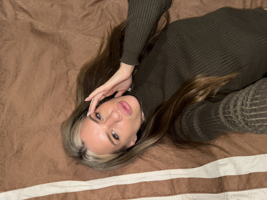 A woman with long, highlighted hair lies on a patterned brown and white bed. She wears a dark green, ribbed turtleneck sweater. Her head is tilted, her eyes looking up at the camera, and her hand gently rests on her forehead. Her lips appear full and slightly glossy. The angle of the shot is from above, focusing on her face and upper body.