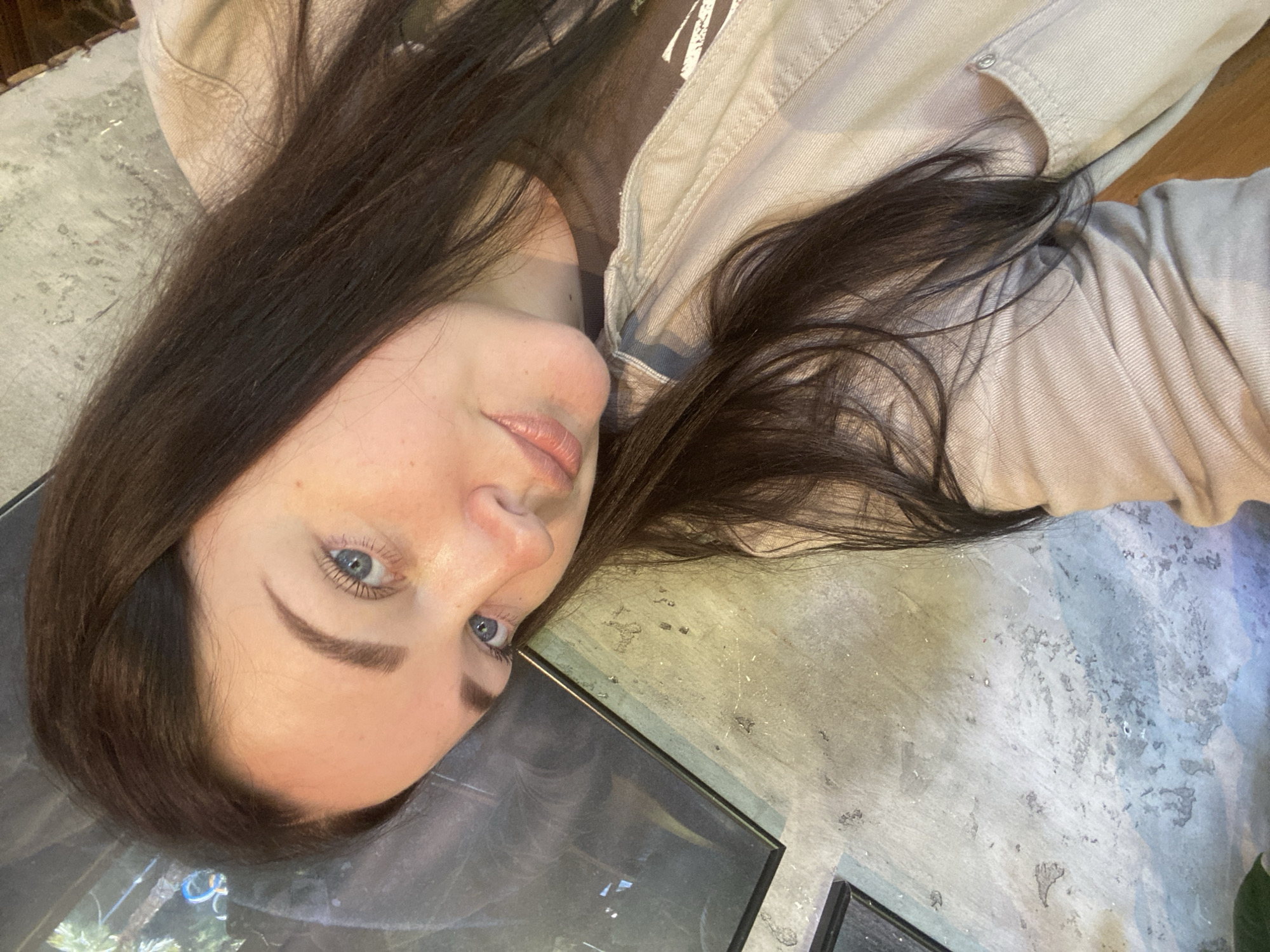 A woman with dark hair and blue eyes is lying on a concrete surface. She is wearing a tan jacket. A framed picture is visible near her head. The location is unknown, and there is no indication it is in Sochi, Russia.