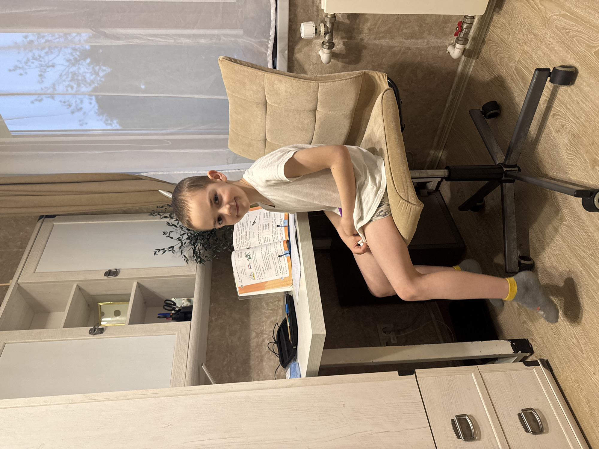 A young boy is sitting sideways in an office chair at a desk in an indoor setting. He wears a white t-shirt and shorts, and gray socks with yellow trim. An open book lies on the desk in front of him. The room contains a built-in cabinet, a window with white curtains, and a radiator. The image appears to be taken in Раздолье, Russia.