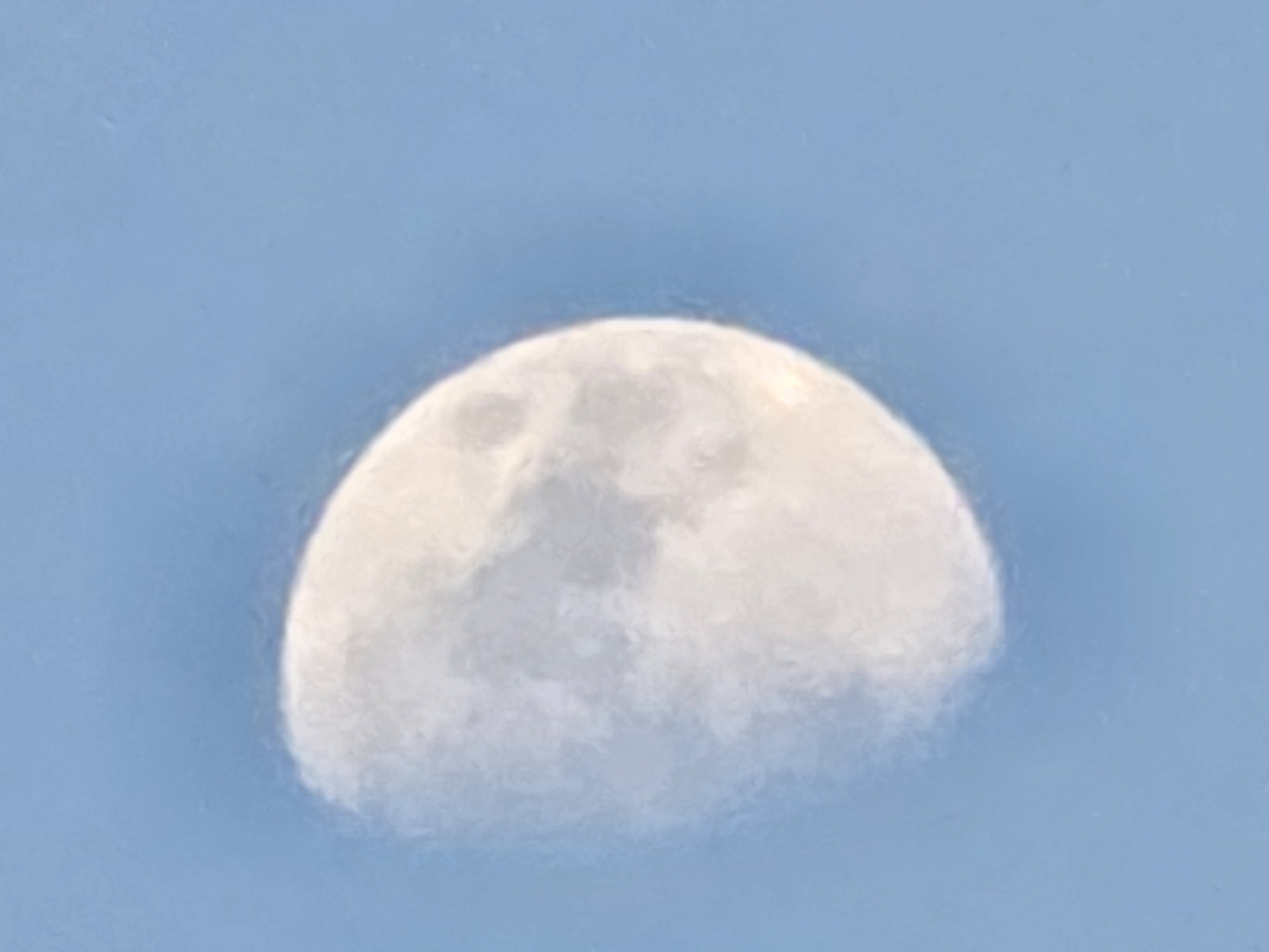 A photograph shows a waxing gibbous moon against a clear blue sky. The moon occupies the left side of the frame, with its bright, illuminated portion visible. The right side of the moon is in shadow, creating a crescent shape. The surface of the moon shows some texture and craters. The sky is a uniform light blue color, devoid of clouds or other celestial bodies. The image appears to be taken from an outdoor location, likely during daylight hours given the brightness of the sky and the moon's visibility. There is no information in the image to suggest a specific location within Yambol, Bulgaria.