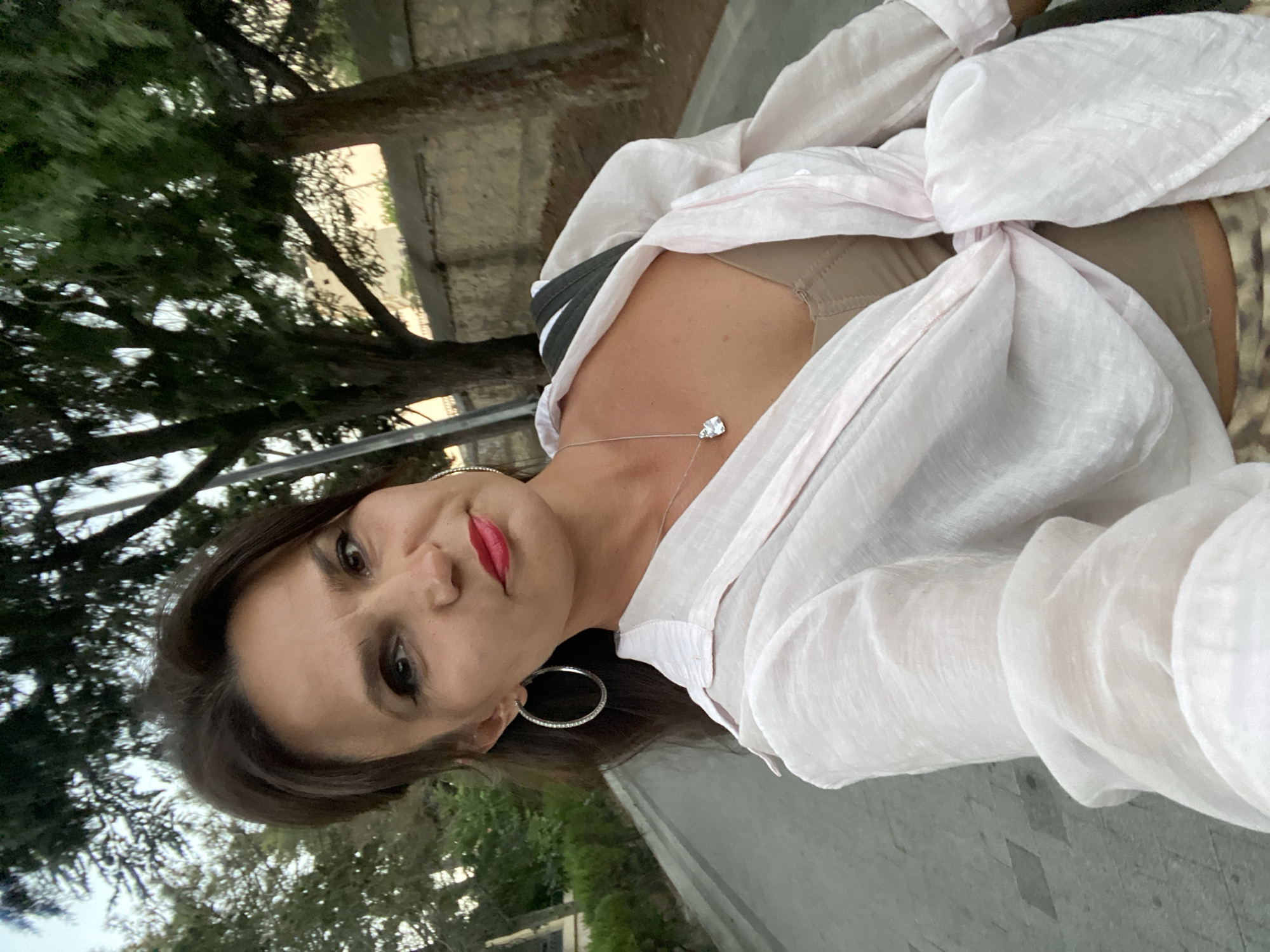 A woman is taking a selfie outdoors in Feodosia, Ukraine. She has dark hair and is wearing a light pink unbuttoned shirt tied at the waist, a beige top, and patterned pants. She is also wearing large hoop earrings and a silver necklace with a pendant. Behind her, there is a paved walkway, trees, and a stone wall. The lighting suggests it is daytime, possibly late afternoon or early evening.