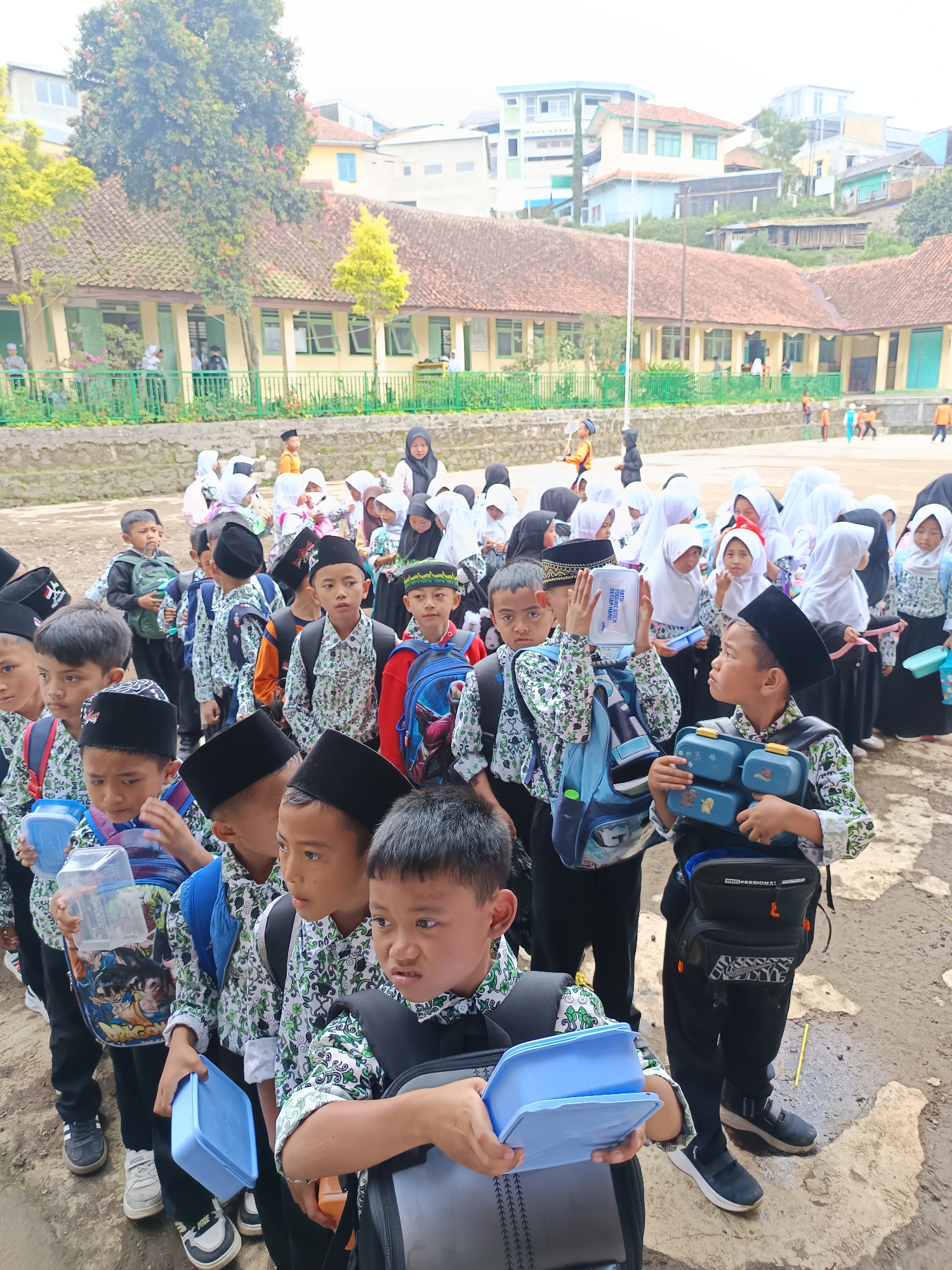 In Gunung Putri, Indonesia, a large group of children, mostly boys, are lined up outdoors. Many of the boys are wearing black caps and uniforms with a green and white pattern. Most children are carrying lunch boxes, predominantly blue. Behind the boys, girls are lined up wearing white hijabs. The backdrop includes a long, low building with a red-tiled roof, suggesting a school. In the distance, there are buildings on a hillside. The ground is a dirt surface.