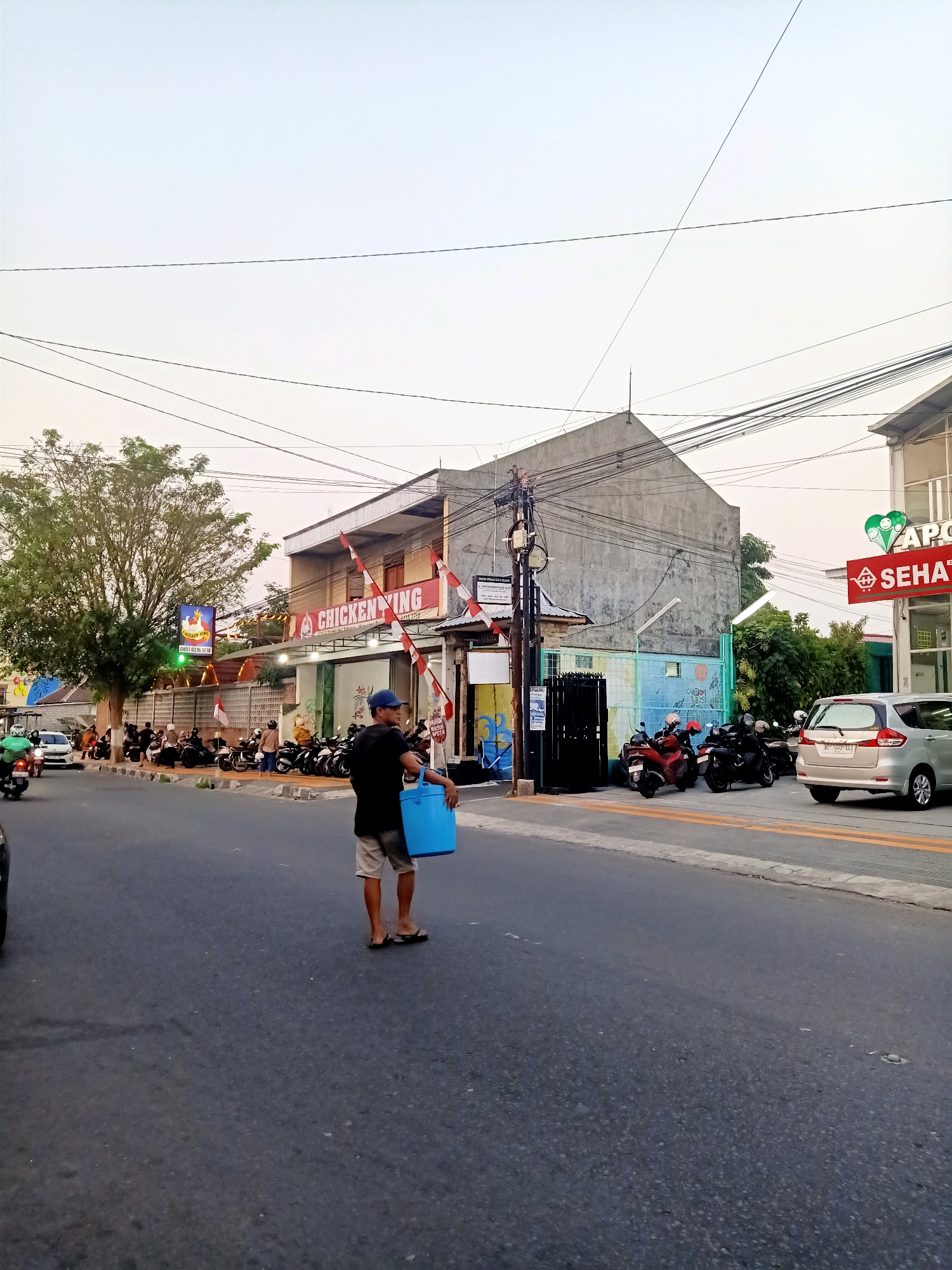 The image depicts a street scene in Klaten, Indonesia, likely in the late afternoon or early evening due to the soft lighting. In the foreground, a man wearing a blue cap, black t-shirt, and light-colored shorts is walking away from the camera, carrying a large, bright blue plastic container in his right hand. He is on the asphalt road, facing towards the right of the frame.
The middle ground features a row of buildings lining the street. The building on the left appears to be a "CHICKEN KING" restaurant, with a sign featuring a cartoon chicken and Indonesian text. Several red and white flags are displayed on the facade of this building and the adjacent one. A large tree with green foliage stands to the left of the restaurant. In front of the buildings, a line of parked motorcycles is visible, suggesting a busy commercial area. People are gathered near the motorcycles and the storefronts. A white car is parked on the right side of the street.
The background is dominated by a pale sky with scattered electrical wires crisscrossing overhead. Another building, possibly an "APOTEK SEHAT" (pharmacy), is visible on the far right, with a green heart-shaped logo and red signage. The environment appears to be an urban or semi-urban setting in Indonesia.