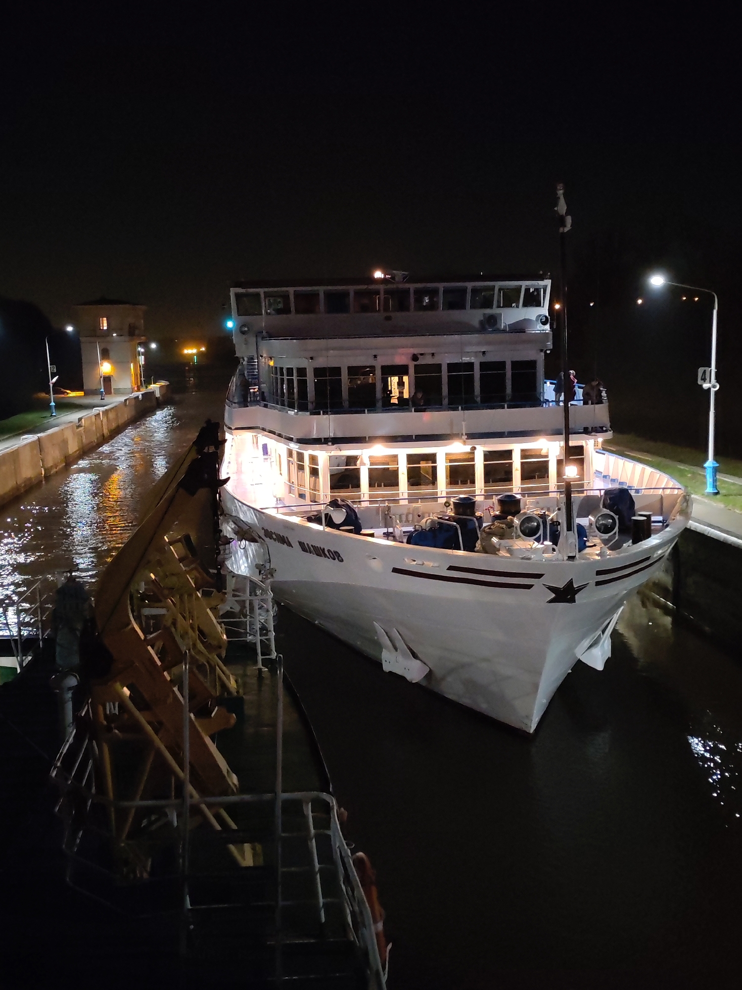 At night in Iksha, Russia, a white passenger ship with multiple decks and illuminated windows is docked at a canal lock. The ship bears the name "ОСИПА ШАПКОв" on its hull, and a black star is painted on the bow. People are visible on the decks. To the left, a smaller boat is alongside the passenger ship, and a metal structure with wooden parts is visible. The canal lock is illuminated, and a building stands at the edge of the waterway. A lamppost is nearby. The water reflects the lights.