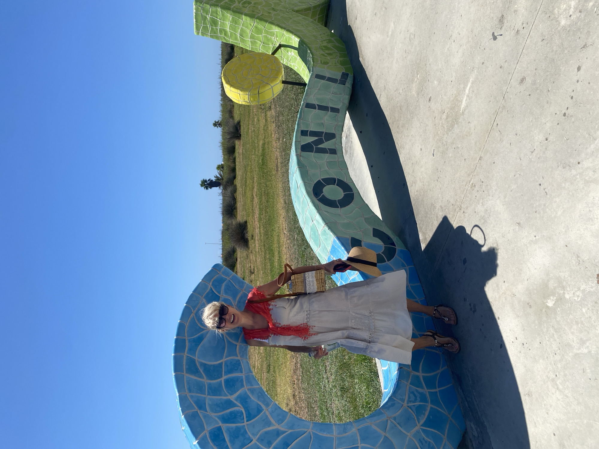 A blonde woman stands in front of a large, brightly colored mosaic sculpture in Conil de la Frontera, Spain. The sculpture appears to be letters spelling out "CONIL" with a circular yellow element on top. The woman is wearing sunglasses, a light-colored tiered dress, a red scarf around her neck, and sandals. She is holding a straw bag over her shoulder and a straw hat in her right hand. The background shows a clear blue sky, green grass, and some sparse vegetation and palm trees. The sculpture is made of blue, green, and yellow tiles. The ground is paved with concrete. The scene is well-lit by sunlight, casting a shadow from the woman.