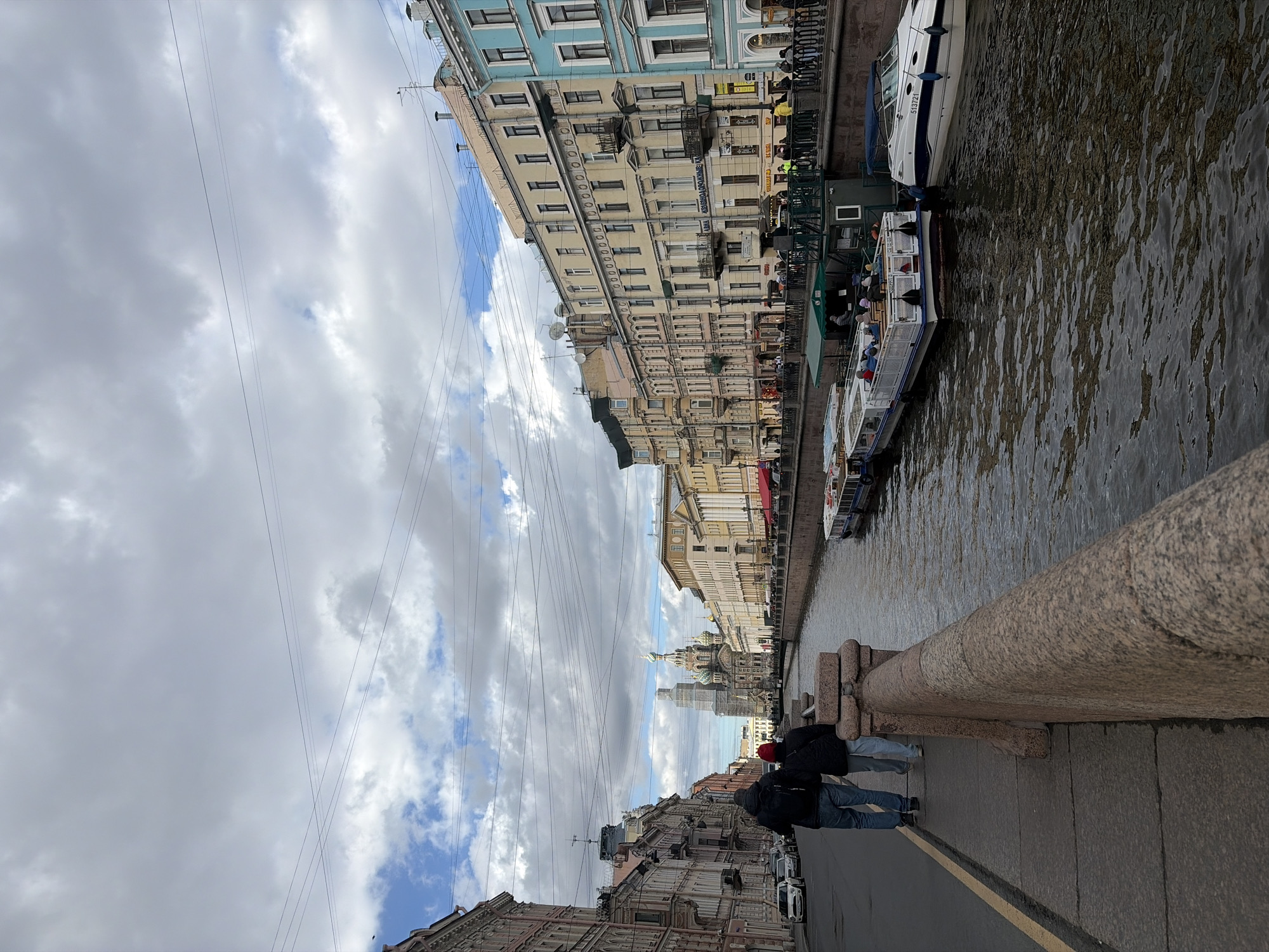 This outdoor image shows a cityscape in Saint Petersburg, Russia, taken from a bridge over a canal. The view is oriented along the canal, showcasing the architectural skyline and the waterway below. The focus is on the perspective leading down the canal, where the iconic spires and domes of the Church of the Savior on Spilled Blood are visible in the distance.
The canal is lined with buildings featuring traditional Russian architecture, including detailed facades and varying heights. There are several boats docked or moving along the canal. Power lines are strung across the sky, adding a modern element to the historical setting.
In the foreground, two individuals stand on the bridge, looking towards the view. The bridge's granite railing is prominently featured. The sky is overcast with clouds, suggesting a slightly overcast day. The image captures the blend of historical architecture and contemporary life in Saint Petersburg.