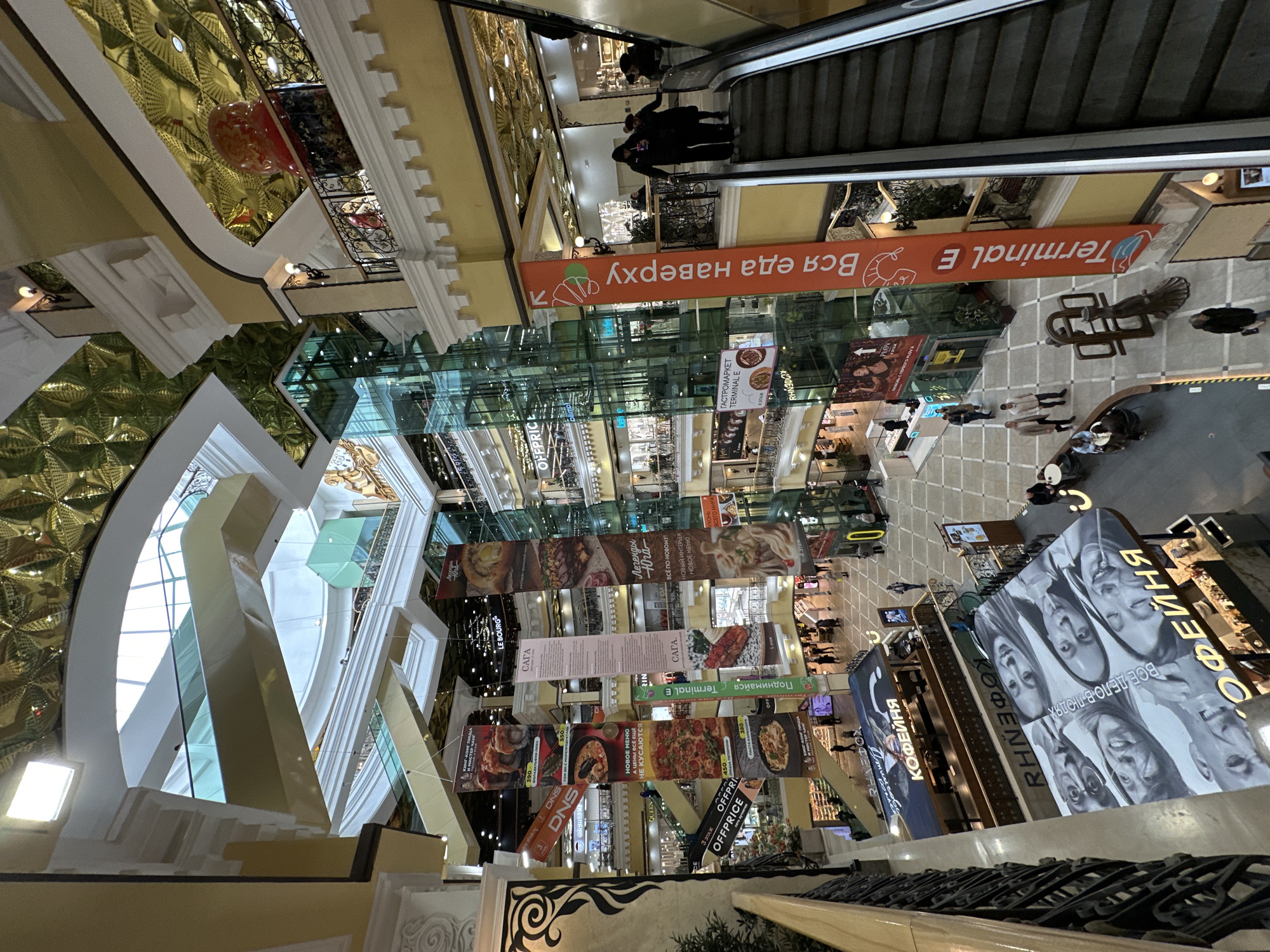 The image presents a high-angle view looking down into the multi-story interior of a busy shopping mall or gastromarket in Yekaterinburg, Russia. The setting combines modern glass architecture with opulent, classical-inspired design elements, including a golden patterned ceiling, ornate black railings, and white balustrades.
Various people are visible across different levels, walking, shopping, and utilizing the escalators. The scene depicts a typical commercial leisure activity, with individuals moving about the space.
Notable details include the intricate ceiling design and the multiple layers of glass and steel creating a spacious atrium. Numerous banners and signs are suspended overhead, predominantly in Cyrillic, advertising food establishments and providing directional information. Visible text includes "Вся еда наверху" (All food upstairs) and "Поднимайся Terminal E" (Go up to Terminal E), indicating a food court named "Gastromarket Terminal E" on an upper floor. Other shop names like "Saga," "Le Bourg," "Kofeynya" (Coffee shop), "OFFPRICE," and "DNS" are present. Food advertisements display dishes with prices, and slogans such as "Легенды Юга" (Legends of the South) and "Новое меню, а цены всё ещё не кусаются" (New menu, and prices still don't bite) are seen. A large black-and-white sign features faces and the text "ВСЕ ДЕЛО В ЛЮД" (It's all about people). A sign indicates "3 этаж OFFPRICE" (3rd floor OFFPRICE), suggesting at least three retail levels. The lighting appears to be a mix of natural light filtering through the glass structures and artificial interior illumination, creating a bright and active atmosphere.