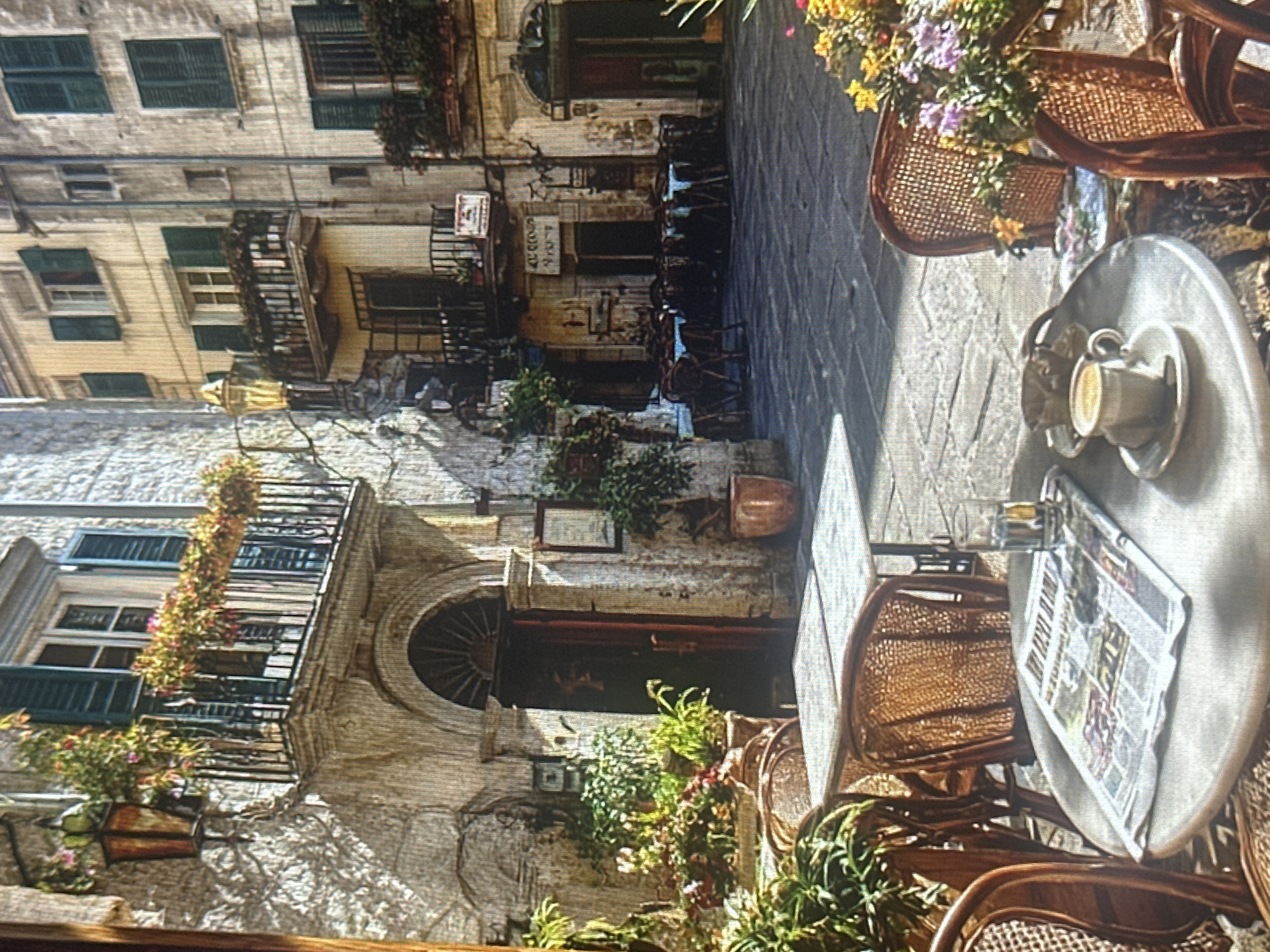 The image depicts an outdoor cafe scene in a European-style alleyway, possibly in Italy. The focus is on a small round table set with a cup of coffee and a newspaper. Wicker chairs surround the table, suggesting a place for patrons to relax and enjoy a meal or drink.
The architecture of the buildings lining the alley is aged and textured, with balconies adorned with overflowing flower boxes. The windows have dark shutters, and the walls show signs of wear, contributing to an old-world charm. Plants and greenery are scattered throughout the scene, adding a touch of life and color.
The lighting suggests it is daytime, with shadows indicating sunlight filtering into the narrow space. The overall atmosphere is peaceful and inviting, evoking a sense of leisurely enjoyment. There is some visible text on the newspaper and signs on the buildings, but it is too blurry to read clearly.