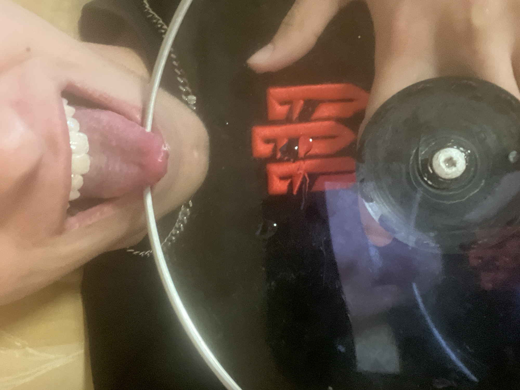 The image shows a human mouth with the tongue extended and touching a white wire. To the right of the mouth, there is a hand holding a black object with a white center. The background is a black surface with a red design and reflections. There is no indication of the city of Sofia, Bulgaria.