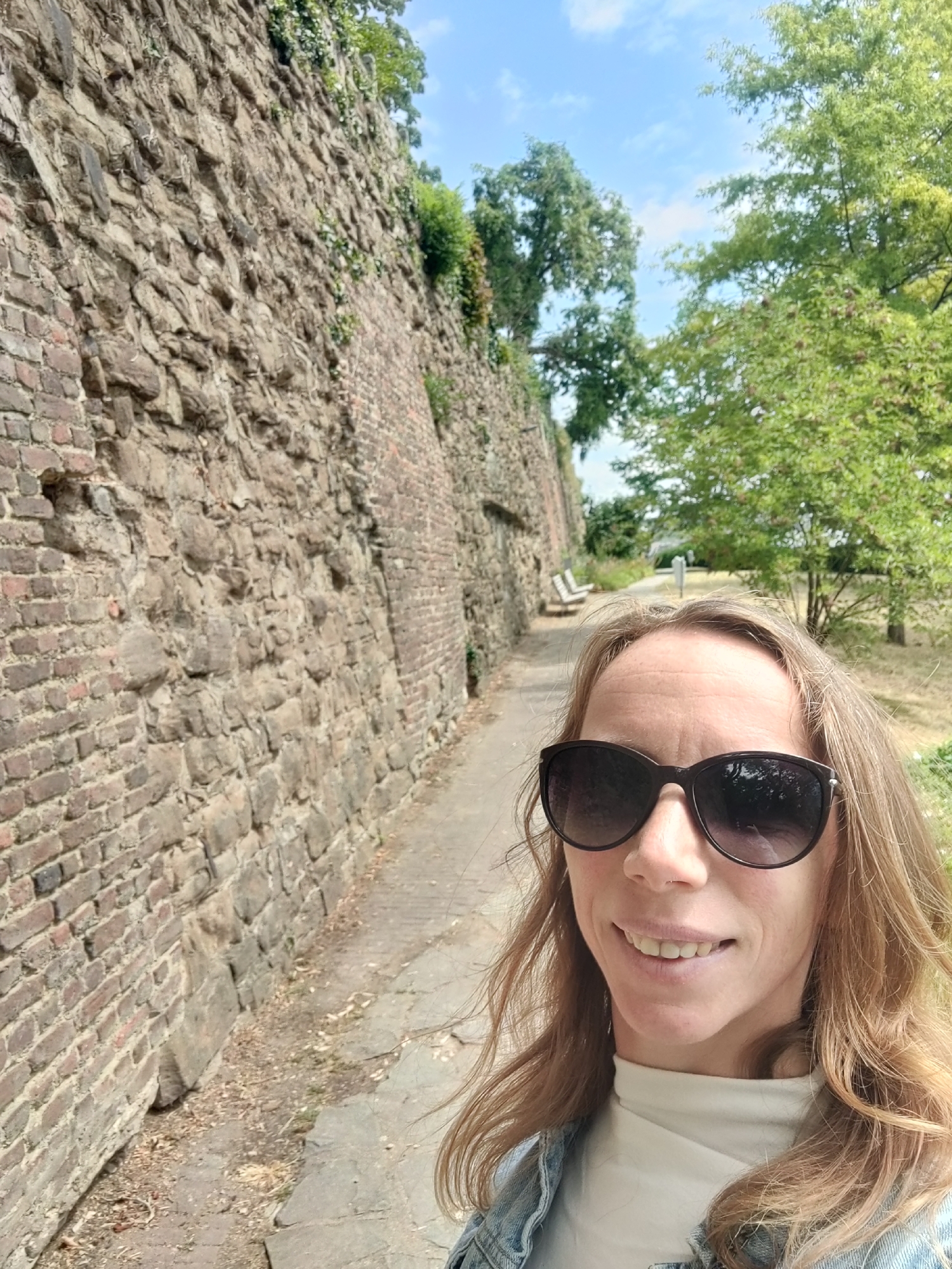A full-body, slightly low-angle, selfie-style photo shows a woman with blonde hair and sunglasses posing in front of an old stone wall. She is wearing a white turtleneck shirt and a denim jacket. The stone wall, which appears to be part of a historical fortification, runs diagonally from the left foreground into the background. The wall is constructed of rough-hewn stones and has some vegetation growing on it. To the right of the wall, a paved pathway runs alongside it, leading into a park-like area with trees and benches visible in the distance. The sky above is blue with some scattered clouds. The scene appears to be outdoors on a sunny day. This location is identified as the Parc de la Citadelle in Liège, Belgium, a historical defense structure.