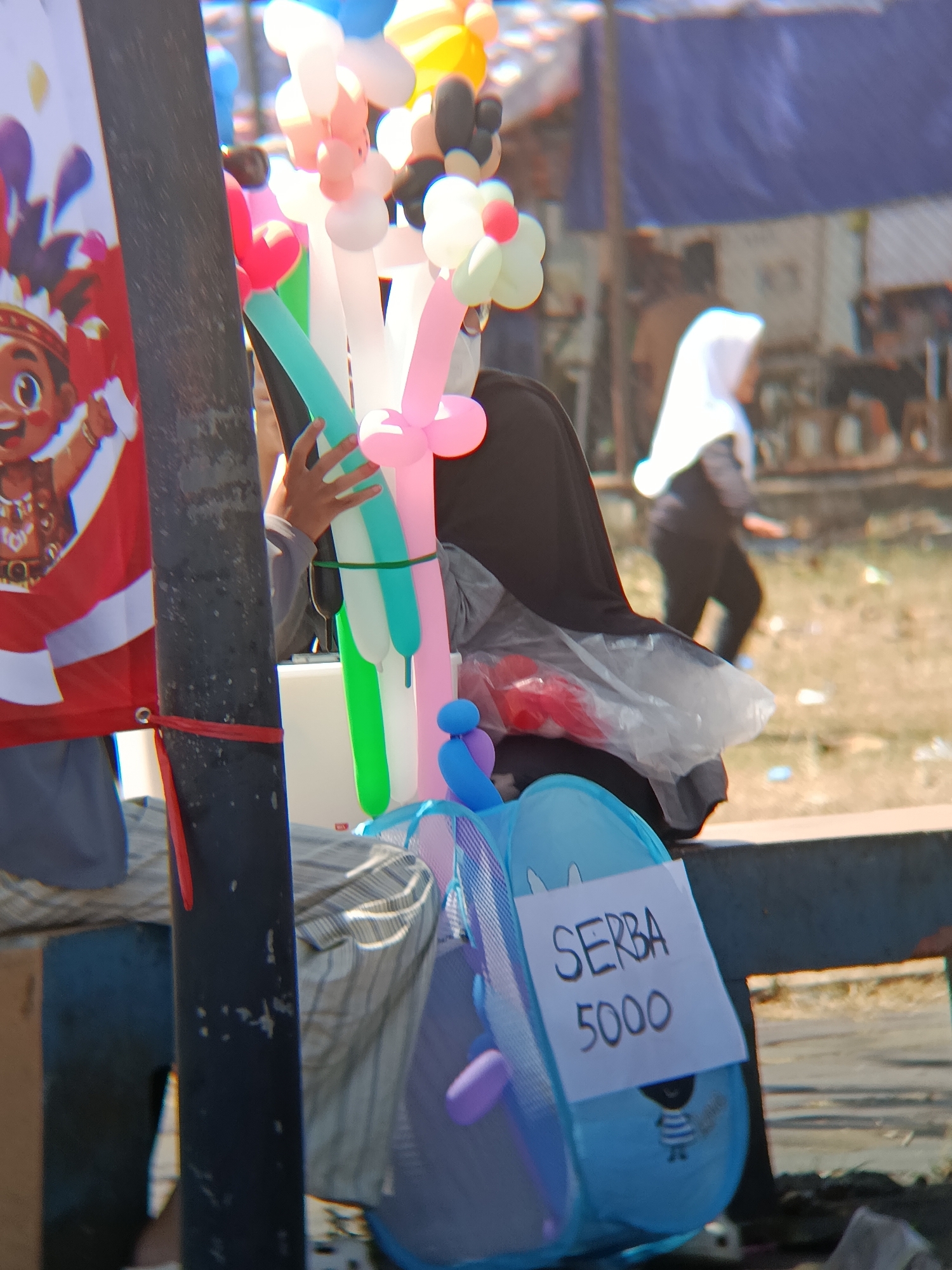 The image depicts an outdoor scene in Desa Kebon Manis, Kecamatan Cilacap Utara, Indonesia, focusing on a stall selling balloons. A person, partially visible and wearing a black head covering, is holding a bunch of colorful, twisted balloons in shades of pink, green, and white. To the left of the balloons, a poster featuring a cartoon character with a headdress is displayed. In the foreground, a blue collapsible basket or toy holder is visible, adorned with a handwritten sign that reads "SERBA 5000", indicating a price. In the background, other individuals are present, one of whom is wearing a white hijab and walking away from the camera. The environment appears to be a public area with tents or tarps set up in the distance and a sandy or dusty ground.