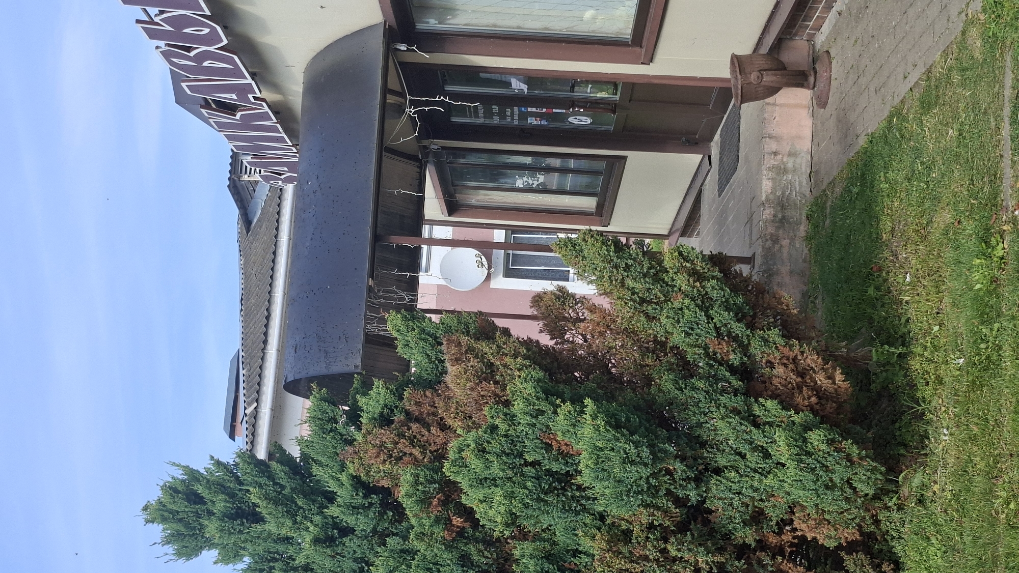 The image shows the exterior of a commercial building with a large green and brown coniferous tree in the foreground. The building has a light beige facade with brown trim and a dark-colored awning over the entrance. The entrance features a double door with windows and a small sign attached to the glass. There are also windows on the side of the building. A satellite dish is mounted on an adjacent building, which has a pinkish hue. The ground is paved with bricks, and there is a patch of green grass to the left of the entrance. A vintage-looking decorative urn is placed on the paved walkway near the entrance. The sky is blue with some light clouds. The sign on the building features large, white letters with a maroon outline that partially reads "АМКАБЬ". This is likely the name of a store or business. Based on available information, the building is located on Vulitsa Pieršamakaj in Myadzyel, Belarus.