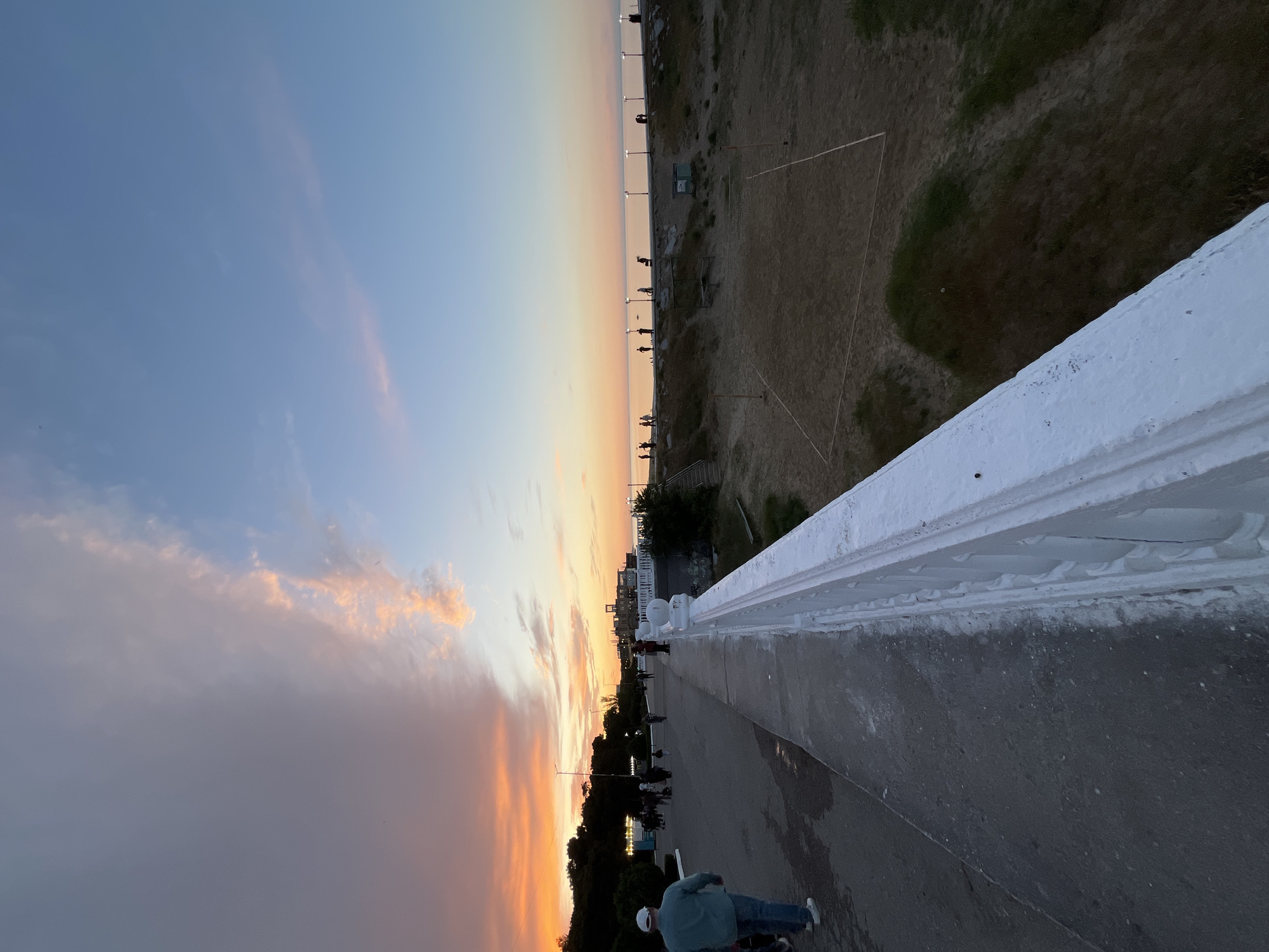 The image captures a scene at sunset in Kaspiysk, Russia. The perspective is from a bridge or elevated walkway overlooking a promenade and the Caspian Sea. A white concrete railing is visible in the foreground, and a sidewalk runs alongside it. People are seen walking and gathering along the promenade below, which is lined with trees and lampposts. A dry grassy area is adjacent to the promenade. The sky is a blend of orange, pink, and blue hues, indicating either sunrise or sunset. A man in a blue jacket and white hat is visible on the sidewalk in the foreground.