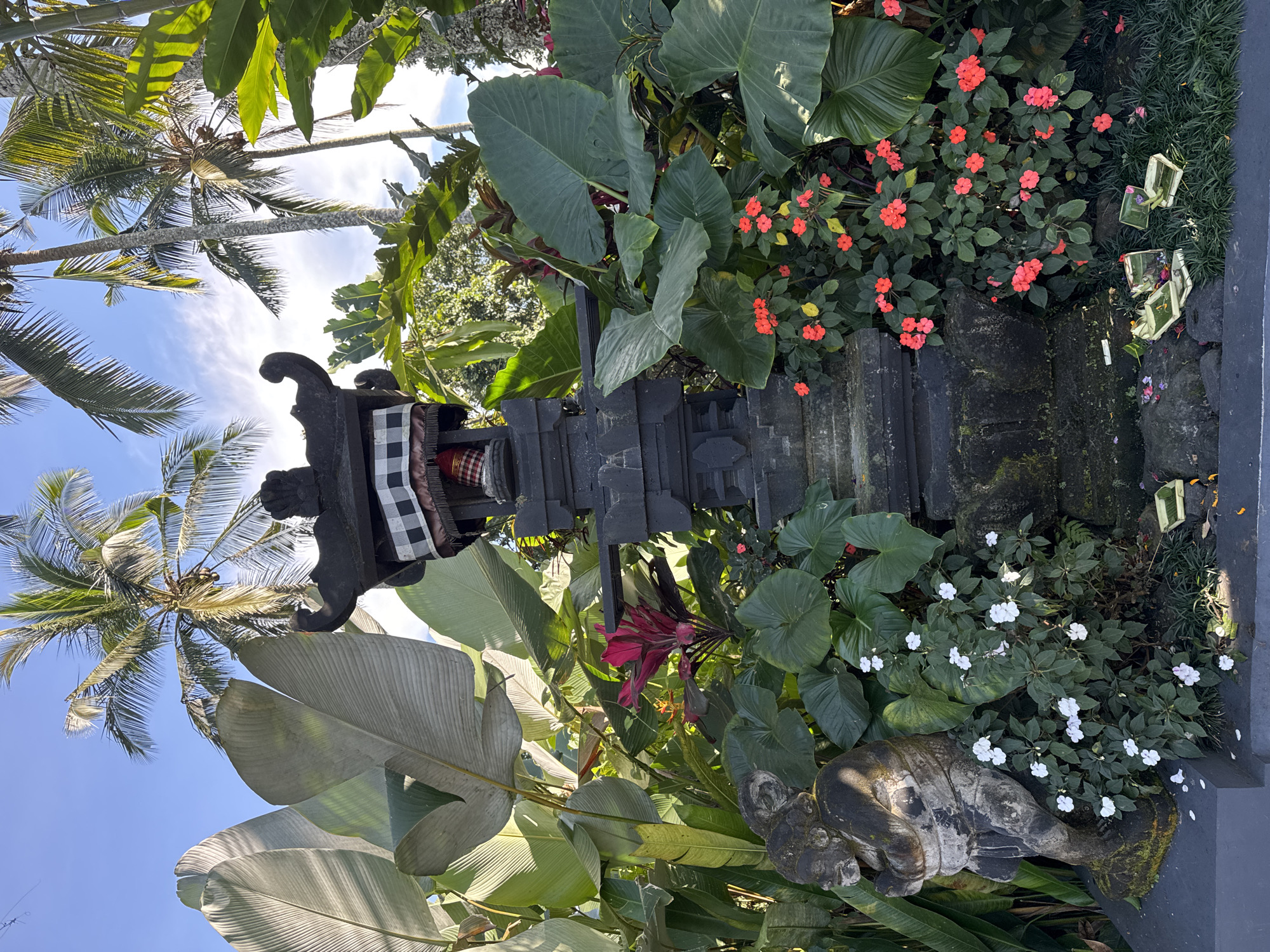 The image captures a traditional Balinese shrine in Rendang, Indonesia. The shrine is constructed of dark stone and adorned with a black and white checkered cloth. Lush tropical vegetation surrounds the shrine, including large green leaves, red and white flowering plants, and palm trees reaching towards the blue sky. A weathered stone statue of a reclining figure is positioned at the base of the shrine. The scene is bathed in natural sunlight.