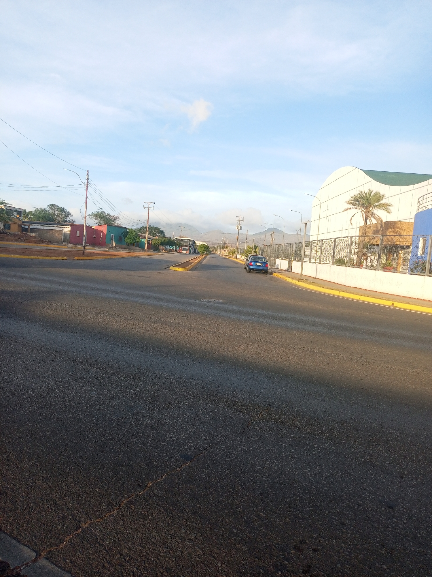 The image depicts a street in Juan Griego, Venezuela. The asphalt road occupies the foreground, stretching into the distance with a clear yellow curb lining the right side. A blue car is parked along the right side of the road, near a white building with a rounded roofline and a fence. A palm tree is near the building.
In the background, mountains are visible. Power poles with overhead lines run along the street. Several buildings are visible on the left side of the road, painted in various colors like red and green. The sky is light blue with some clouds. The light suggests it is either early morning or late afternoon.