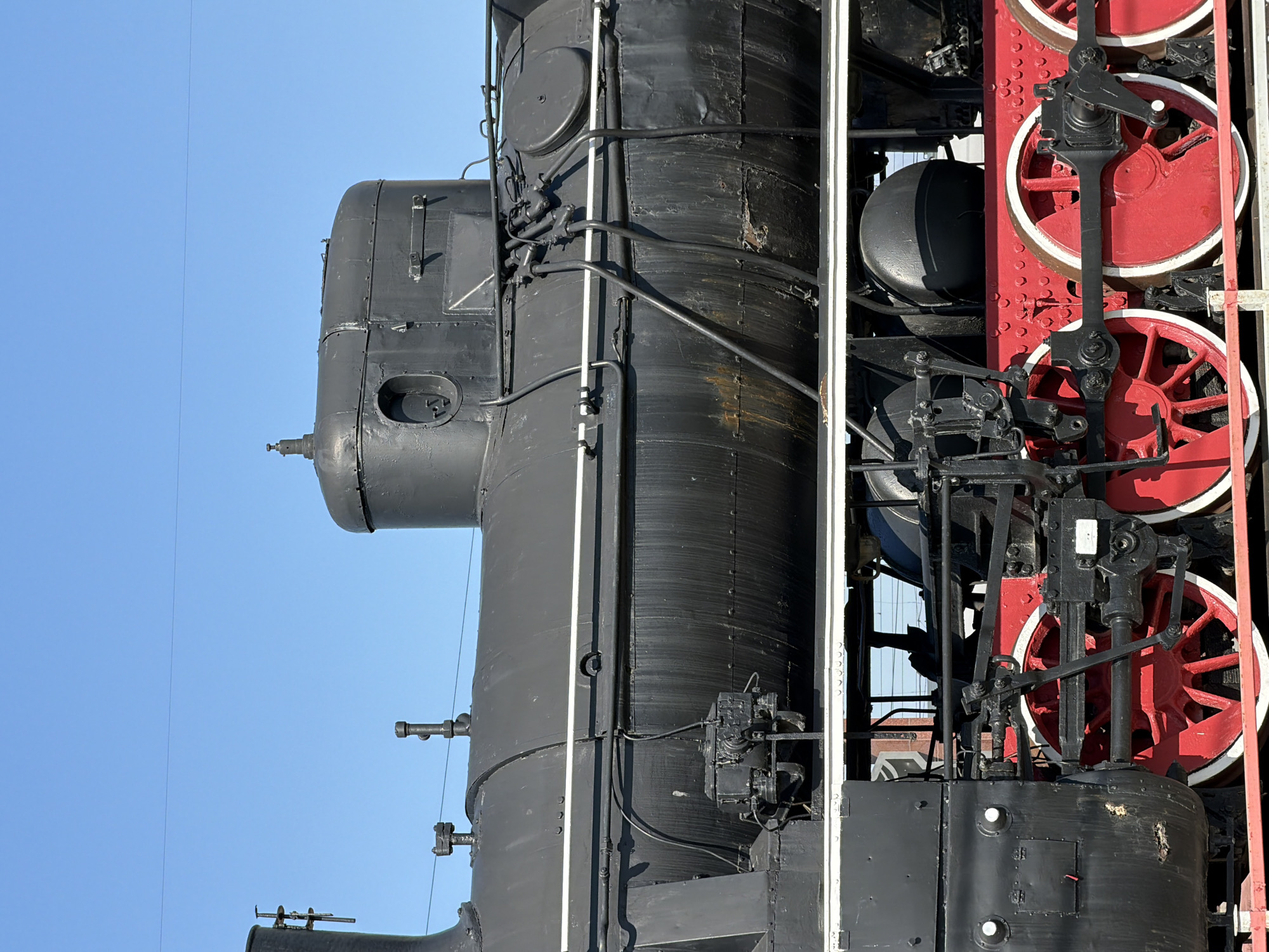 The image shows a detailed view of a black steam locomotive with red wheels, set against a clear blue sky. The shot focuses on the front portion of the engine, displaying its intricate mechanical components, pipes, and gauges. The red wheels contrast sharply with the black body of the locomotive. The location is Petrov Val, Russia, as that's the only information known about the image.