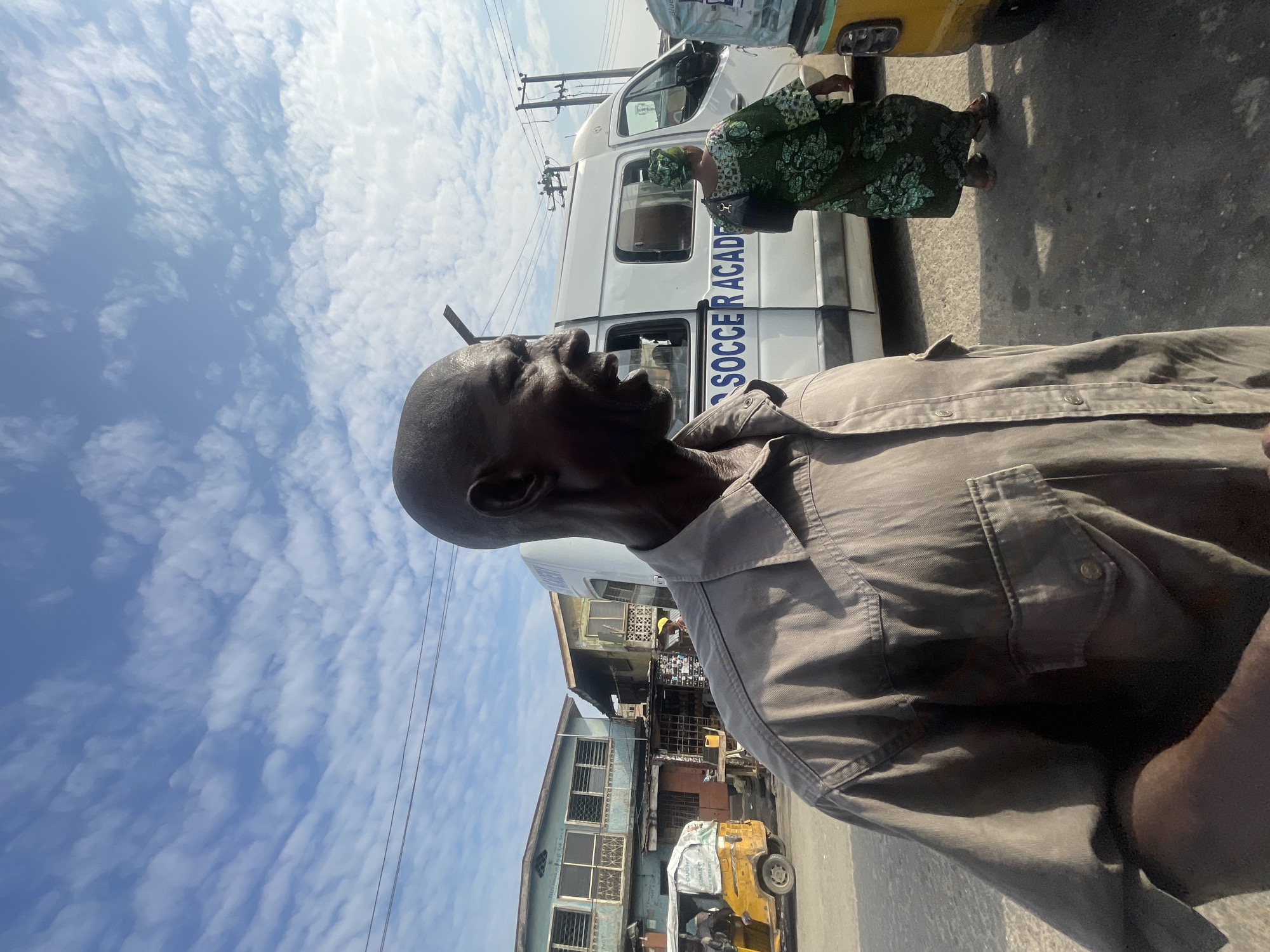 The image captures a street scene in Akowonjo, Nigeria, under a partly cloudy sky. In the foreground, a bald Black man is shown from the chest up, wearing a light brown button-down shirt with two chest pockets. He is looking to his right with his mouth open as if speaking. Behind him, a white van with "SOCCER ACADEMY" written in blue letters on its side is parked. To the left of the van, a yellow auto-rickshaw is visible. Further in the background, several multi-story buildings with windows and balconies line the street. A woman in a green and black patterned dress and headscarf walks away from the van towards the right side of the frame. The ground is paved with asphalt, and power lines are visible crisscrossing the sky.