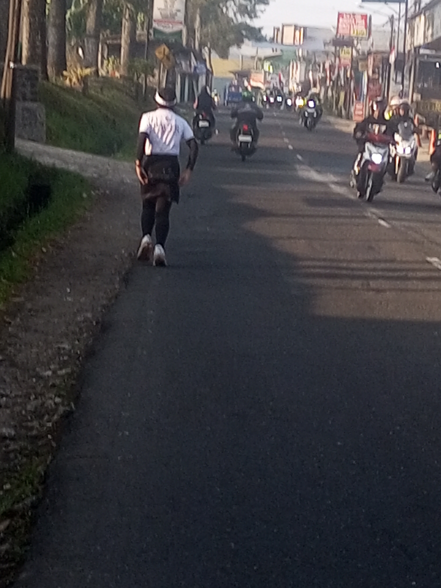The image depicts a man running along a paved road in Garung, Indonesia. He is wearing a white t-shirt, black shorts, black leg warmers, and a white head covering. He is running away from the camera. The road is lined with trees and vegetation on the left, and businesses and buildings on the right. Several motorcycles are visible on the road, some carrying riders, suggesting a procession or event. The lighting suggests it is either early morning or late afternoon.