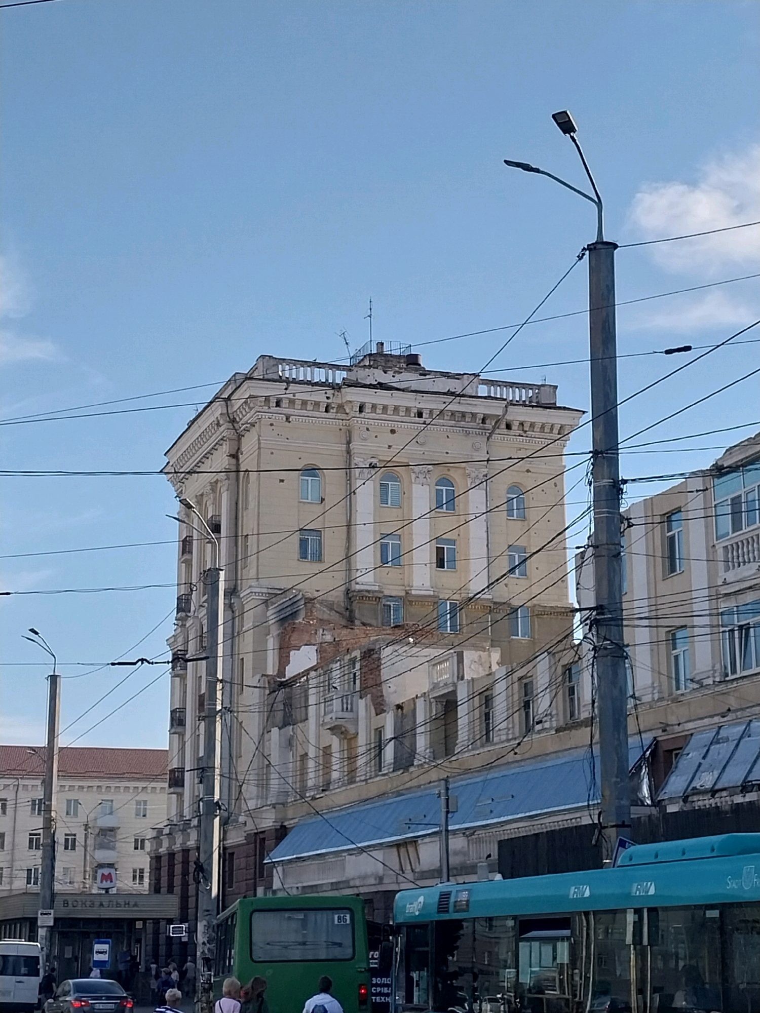 The image captures a street scene in Dnipro, Ukraine, likely near the central train station given the sign reading "ВОКЗАЛЬНА" (VOKZALNA, meaning railway station) visible on a building. The main subject is a damaged, pale yellow apartment building with several stories, exhibiting significant structural damage, including missing facade sections and visible brickwork. The building has arched windows on the upper floors and rectangular windows on lower floors, some with what appear to be white shutters or blinds. The damage suggests it has been impacted by shelling or explosions.
In the foreground, a light blue bus with the branding "Furion" is partially visible, its windows reflecting the surrounding environment. To the left, a green bus with the number "86" displayed on its rear is also present. Several vehicles, including a dark-colored sedan and a white van, are on the street. A crowd of people, some walking, some standing, are visible near the entrance of the "ВОКЗАЛЬНА" building.
Overhead, a complex network of overhead power lines, likely for trolleybuses, crisscrosses the sky, supported by tall grey poles, one of which is prominent in the right-center of the image. Streetlights are also visible on these poles. The sky is a clear, pale blue with some wispy white clouds. The overall impression is one of urban infrastructure, partially intact and partially showing the effects of conflict, with daily life continuing amidst the damage.
