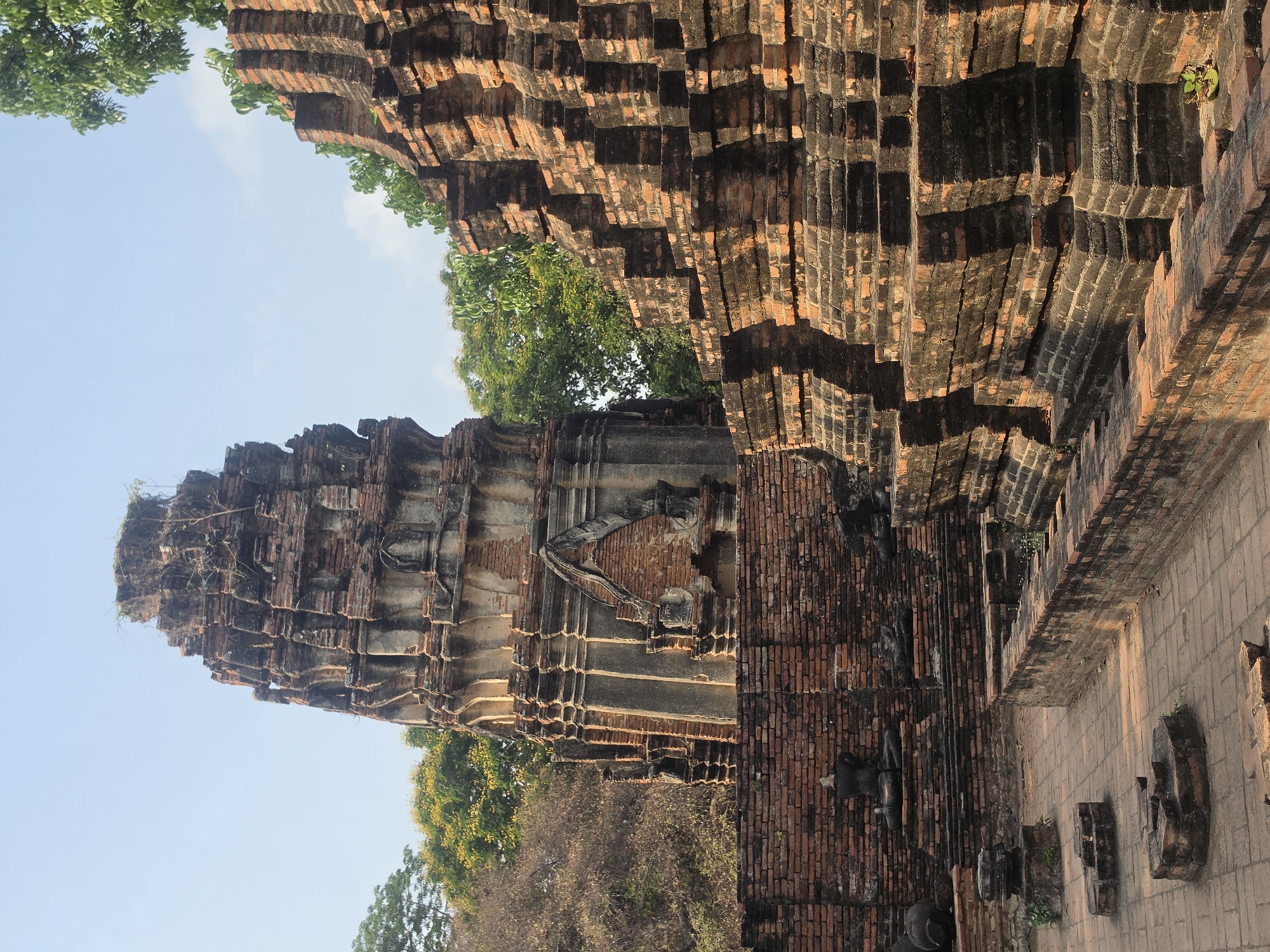 The image depicts an ancient temple ruin in Phra Nakhon Si Ayutthaya City Municipality, Thailand. The ruins are constructed primarily of brick, with sections weathered and broken. The temple structure exhibits multiple levels and a tall central spire. Trees and vegetation grow among the ruins. The sky is partially visible in the background.