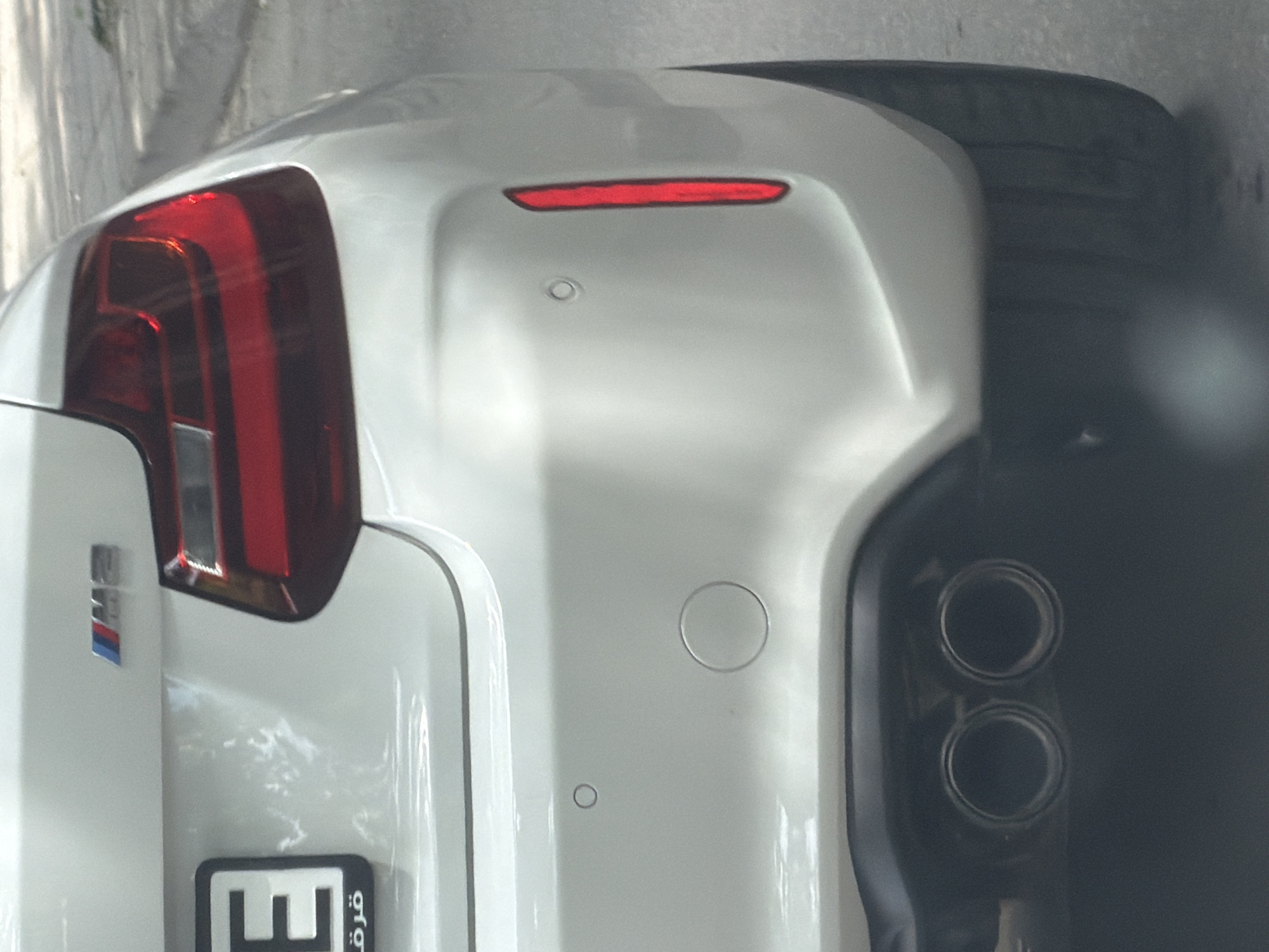 The image shows the rear of a white BMW M car. It includes the taillight, M badge, rear bumper with parking sensors, license plate (likely from the Middle East), and a dual exhaust system. The location is unknown, and there is no indication it is in Plovdiv, Bulgaria.